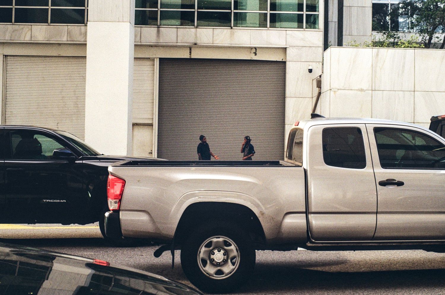 Two people stand near a garage door beside a silver Toyota Tacoma pickup truck parked on a city street.