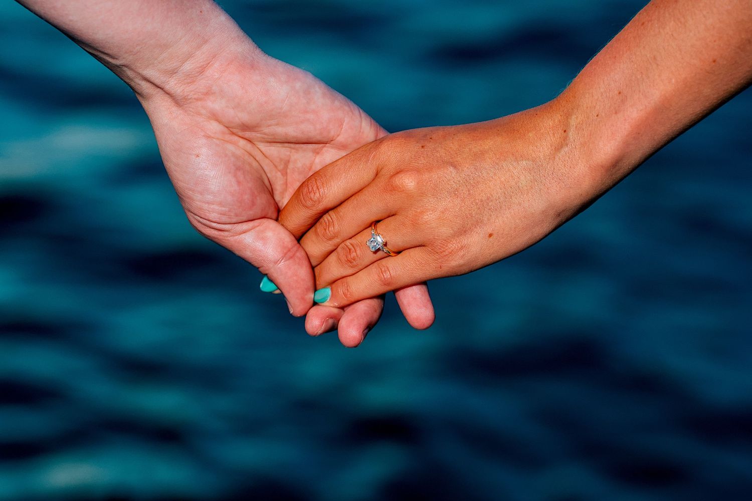 Two hands holding each other over turquoise water, one wearing a diamond engagement ring with teal nail polish.