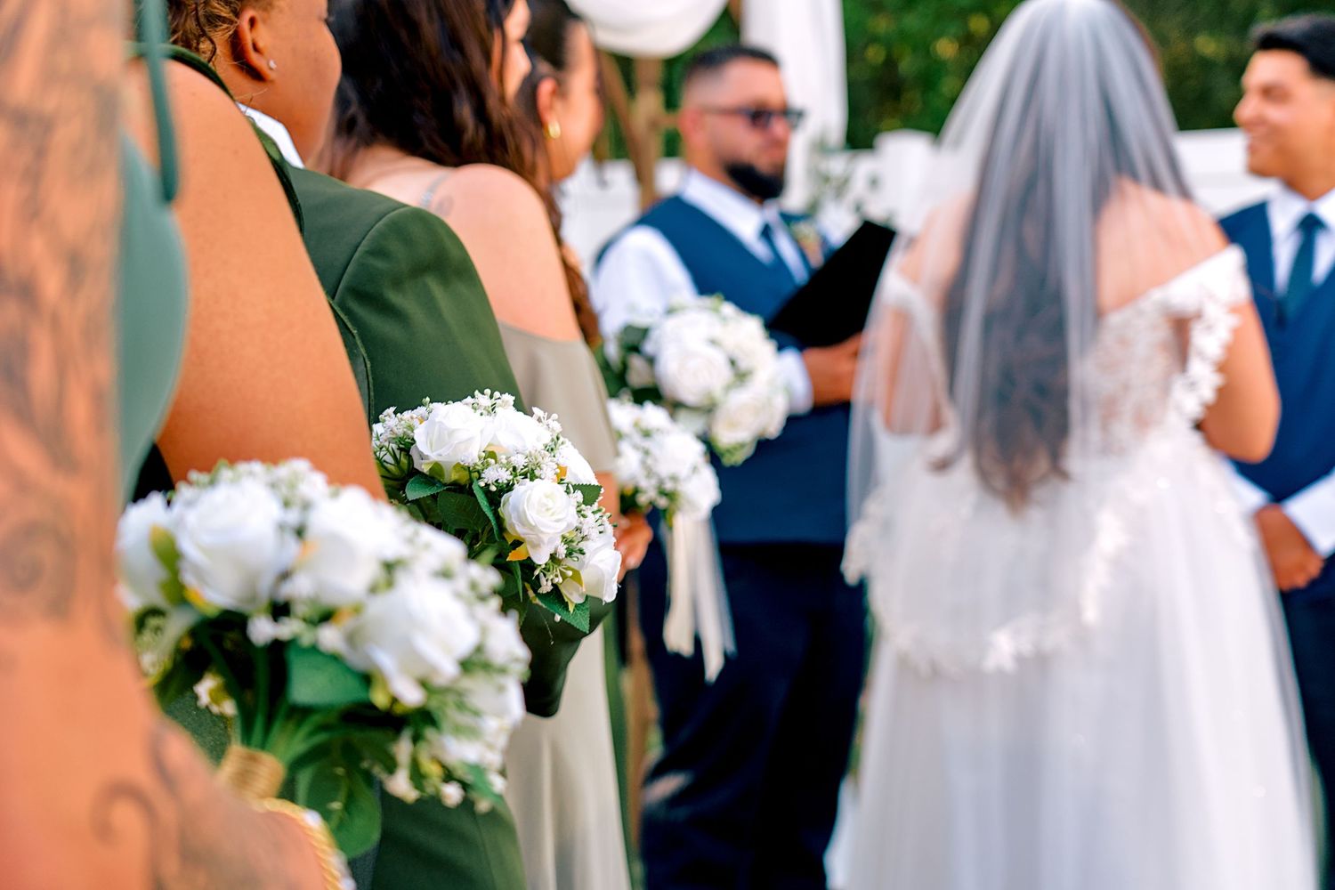 Bridesmaids in sage green dresses holding white bouquets at an outdoor wedding ceremony.