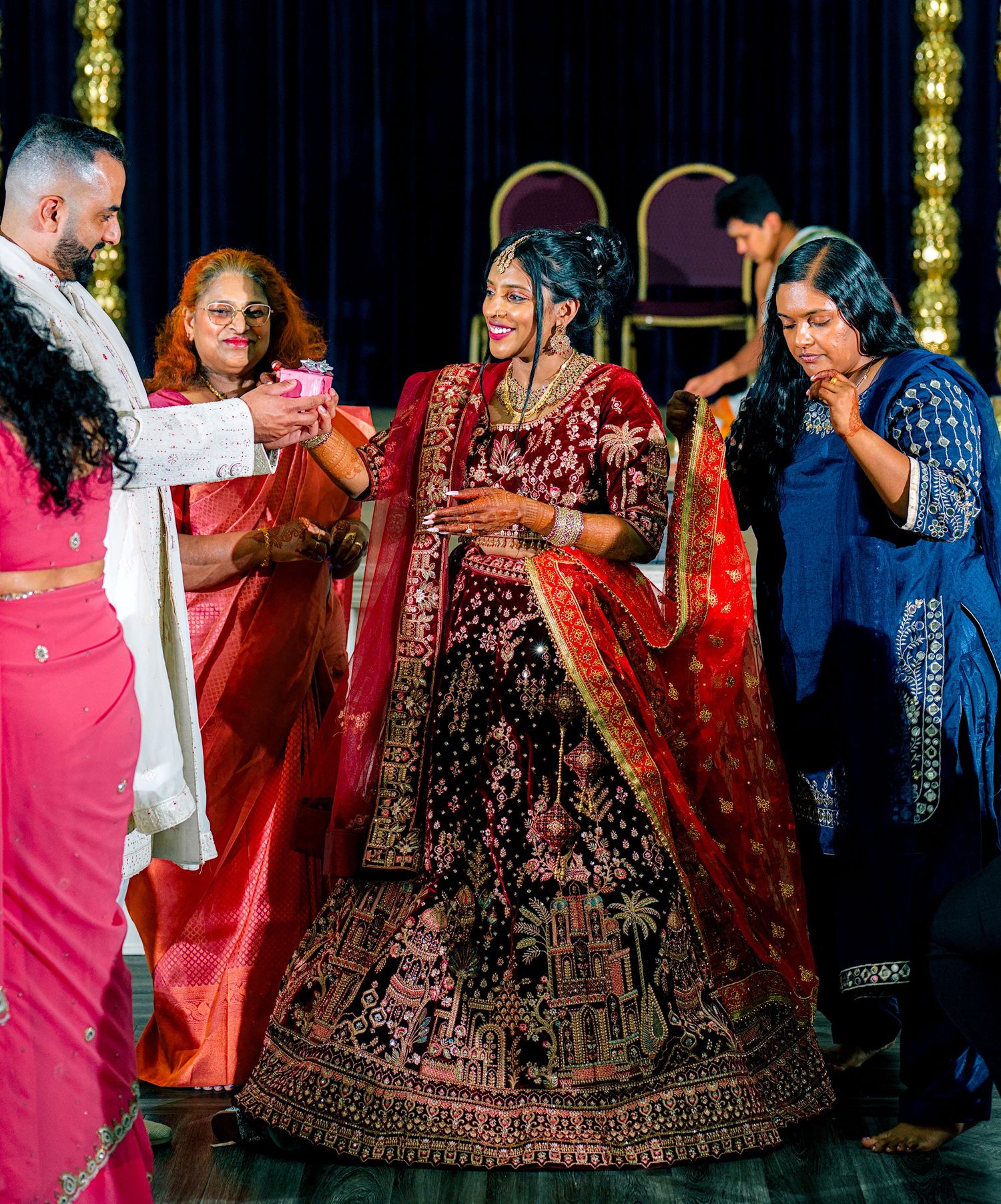 Bride in red bridal lehenga smiling during traditional Indian wedding ceremony with family.