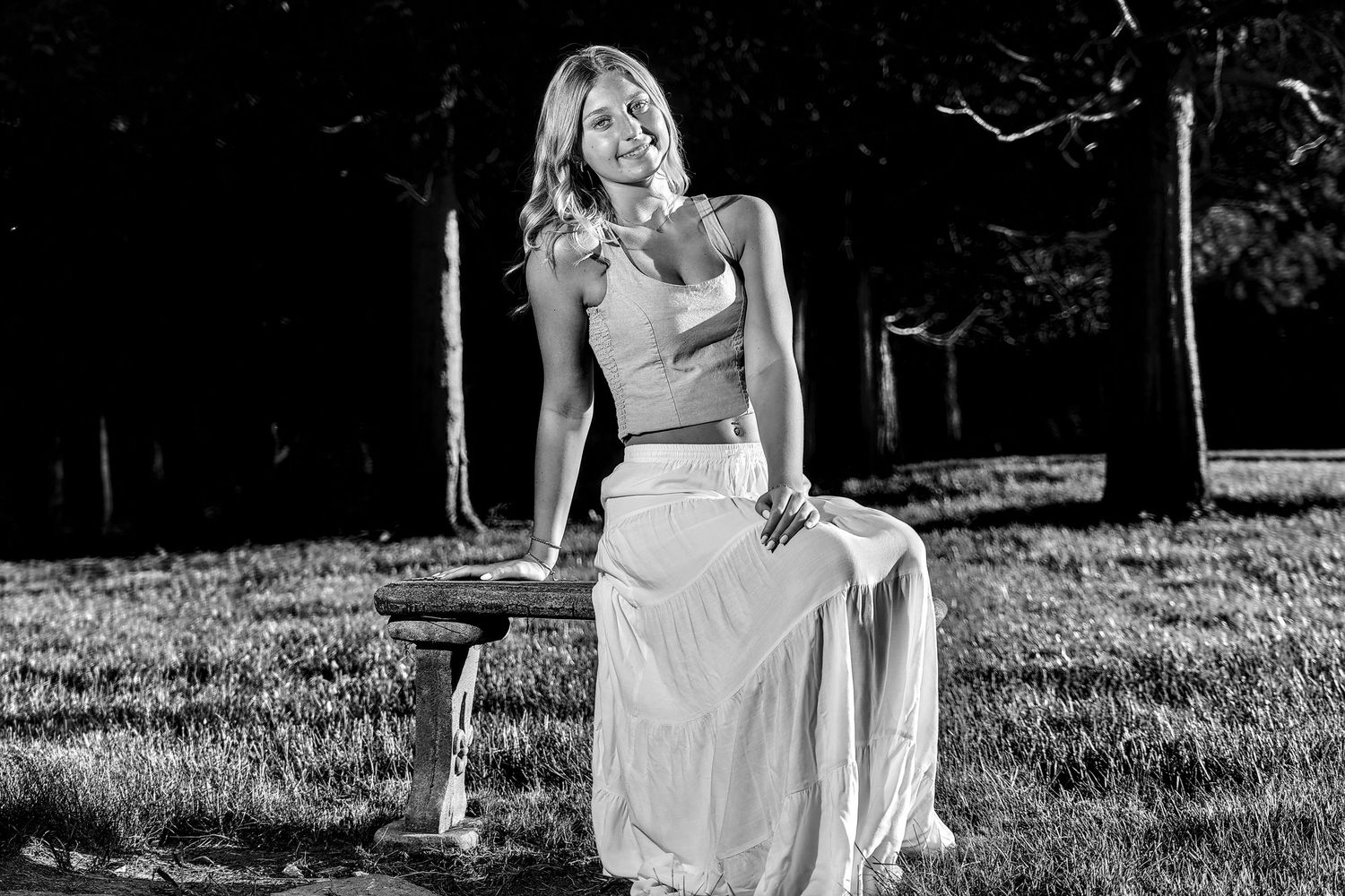 Woman in crop top and flowing skirt poses on bench in black and white outdoor portrait.