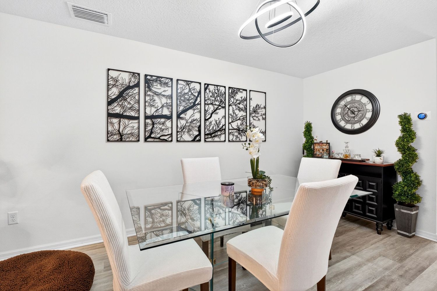 Elegant dining room featuring a glass table, white upholstered chairs, multi-panel tree artwork, and decorative clock on white walls.