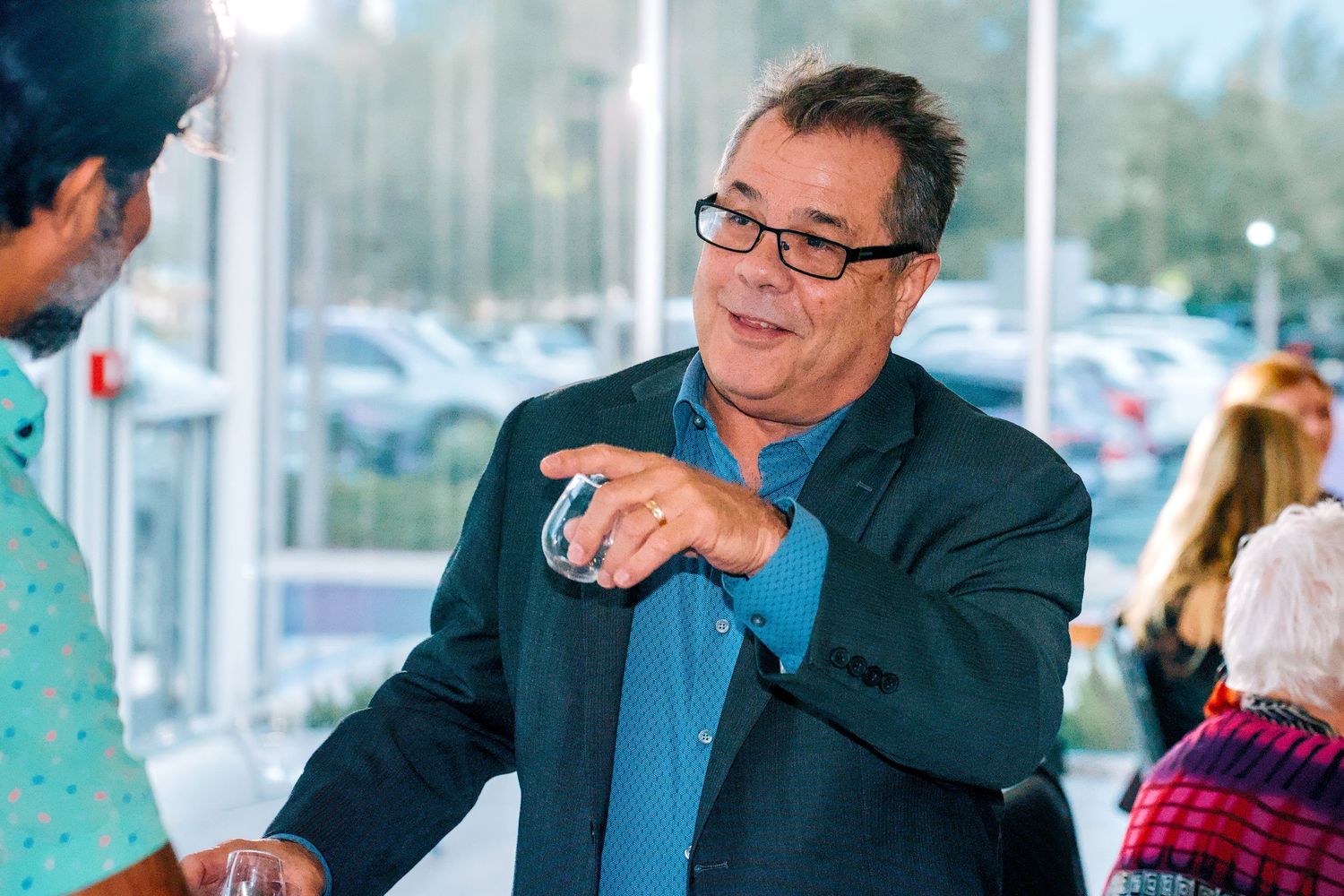 Man in blue shirt and blazer holding a drink glass, smiling and conversing at an outdoor event.