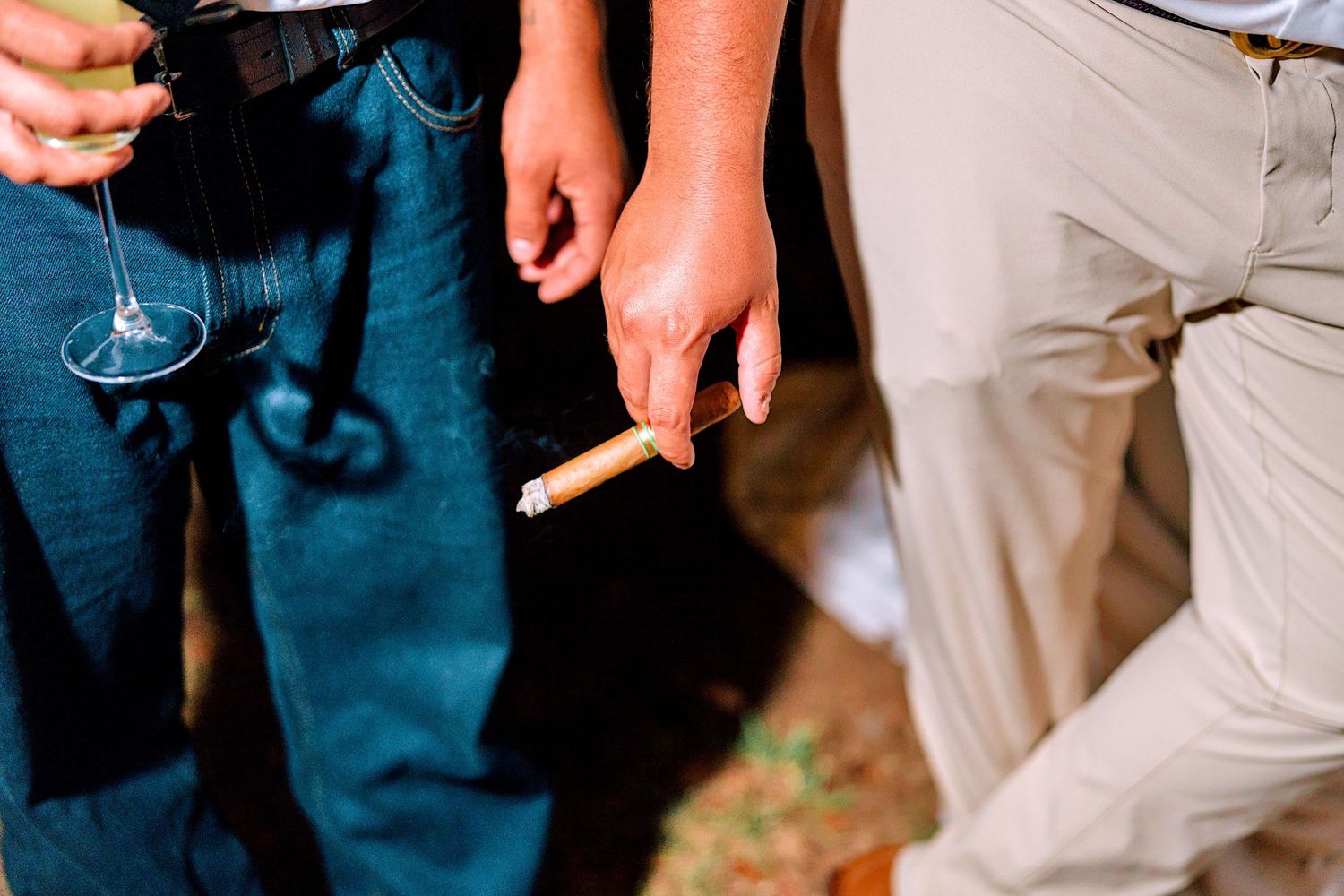 Close-up of hands holding a lit cigarette and wine glass at an outdoor wedding celebration.