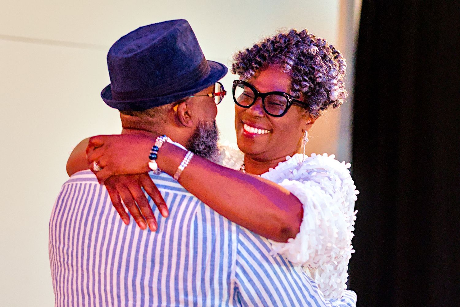 Two people sharing a warm embrace, smiling joyfully, one wearing a blue hat and striped shirt.