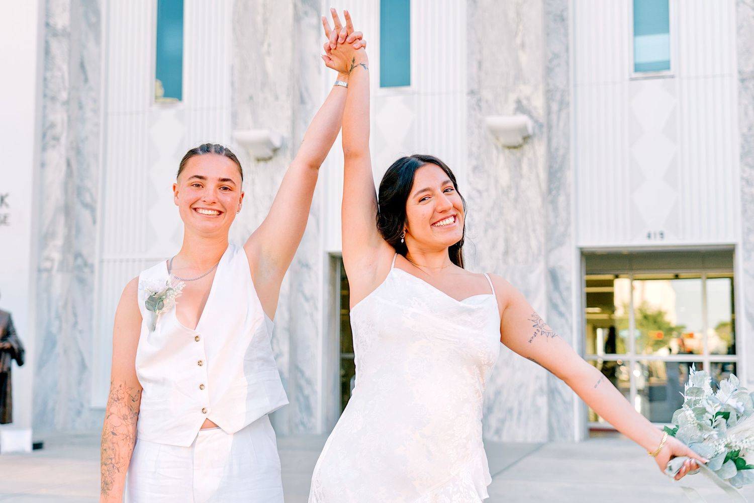 Two brides in white celebrate their wedding day outside a courthouse, raising their joined hands in joy.