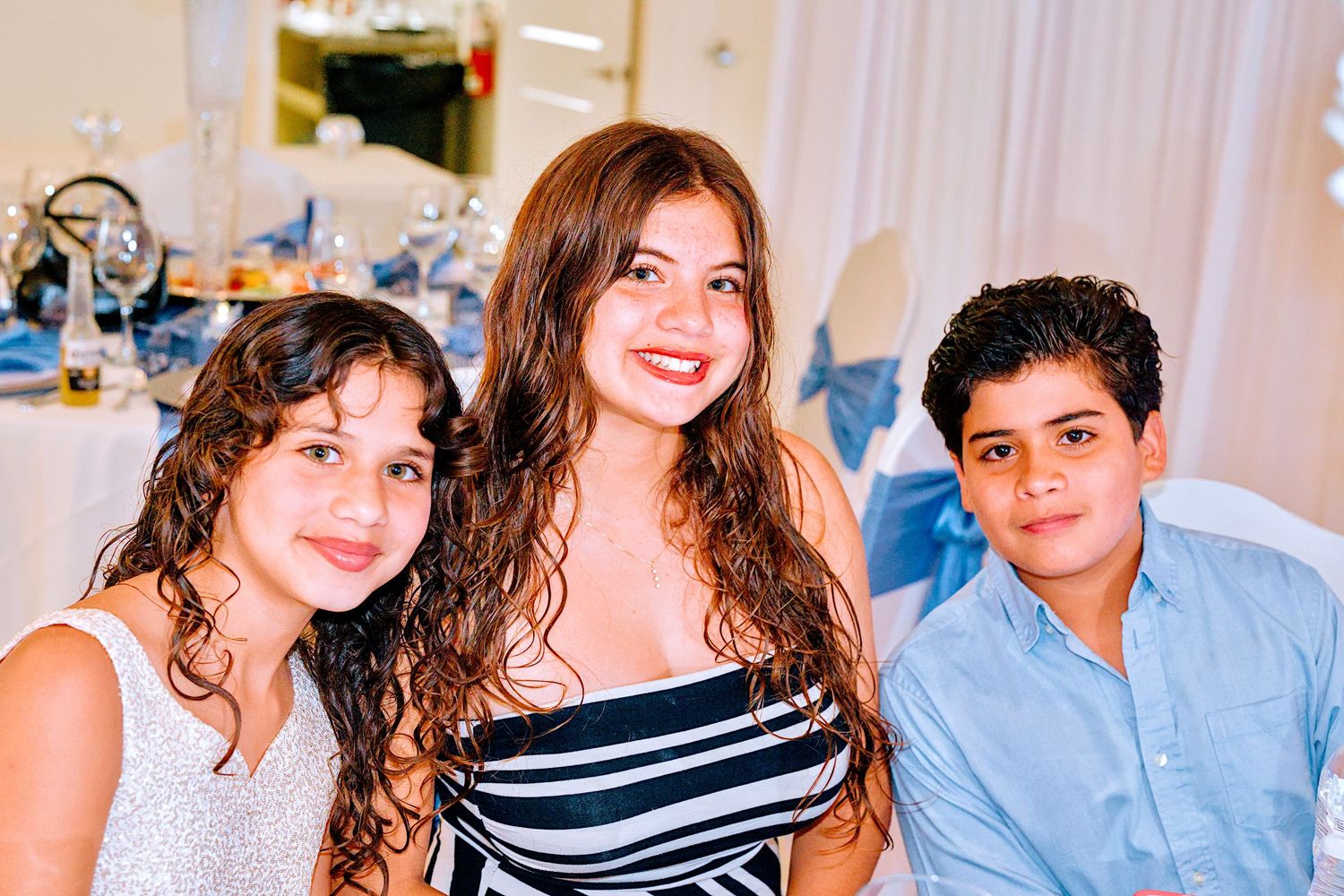 Three smiling siblings posing together at a festive event, two girls and a boy in semi-formal attire.