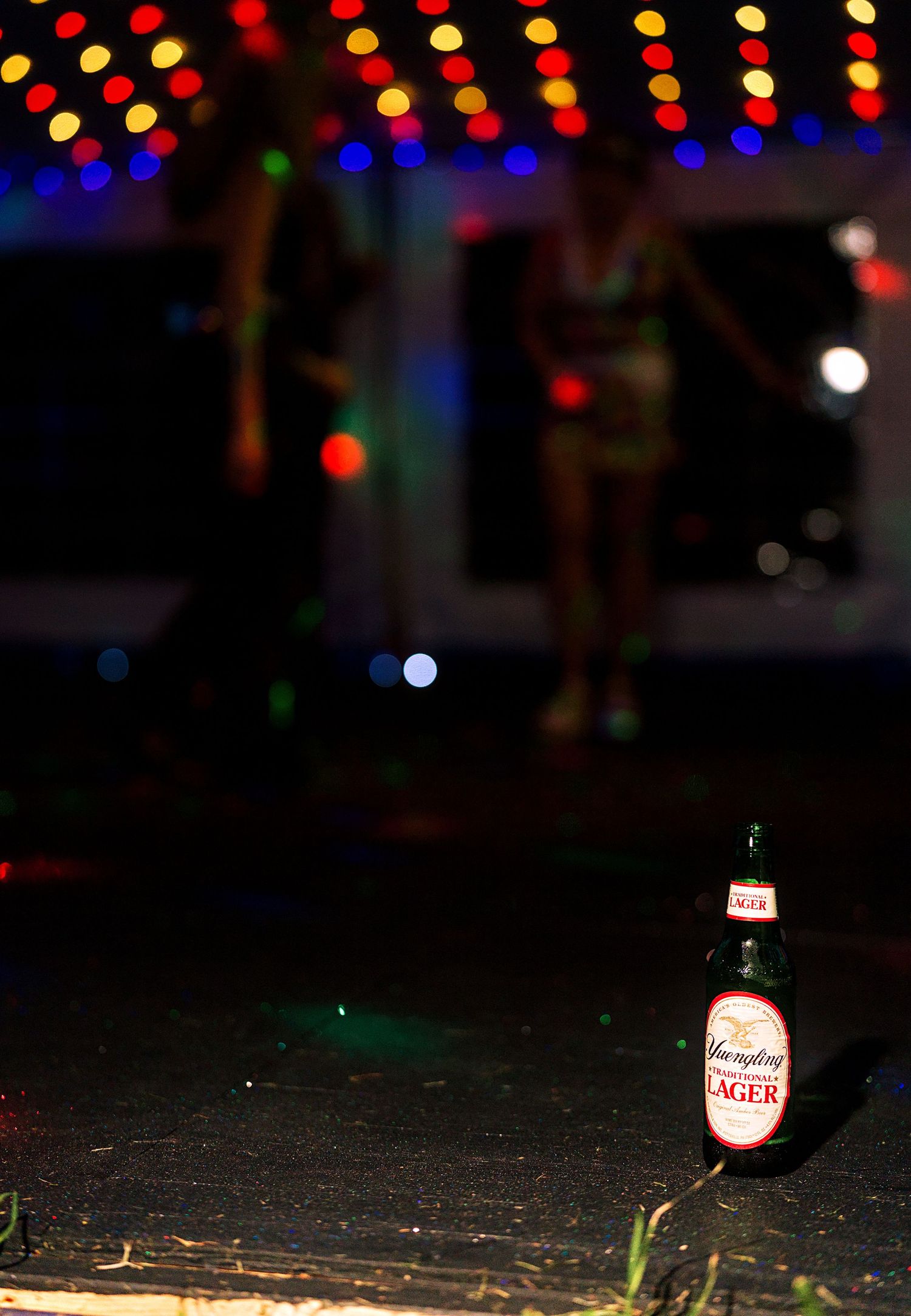A Yuengling Lager beer bottle on the ground at a colorful outdoor disco party with dancers in the background.