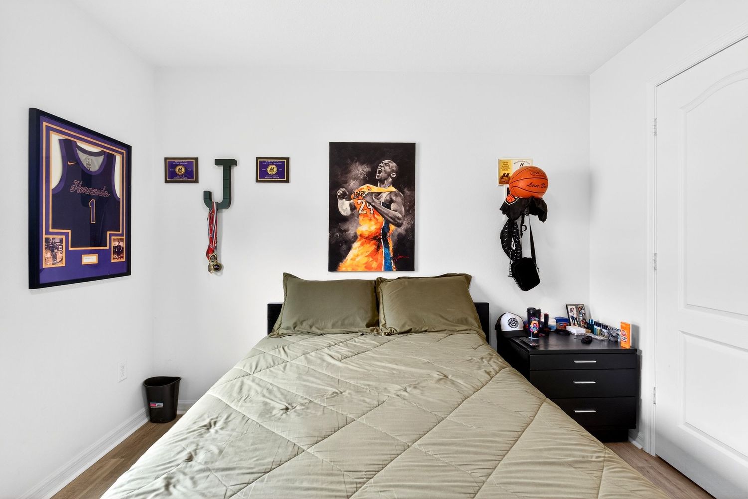 Cozy bedroom with olive green bedding, basketball memorabilia, framed jersey, and sports posters on white walls.