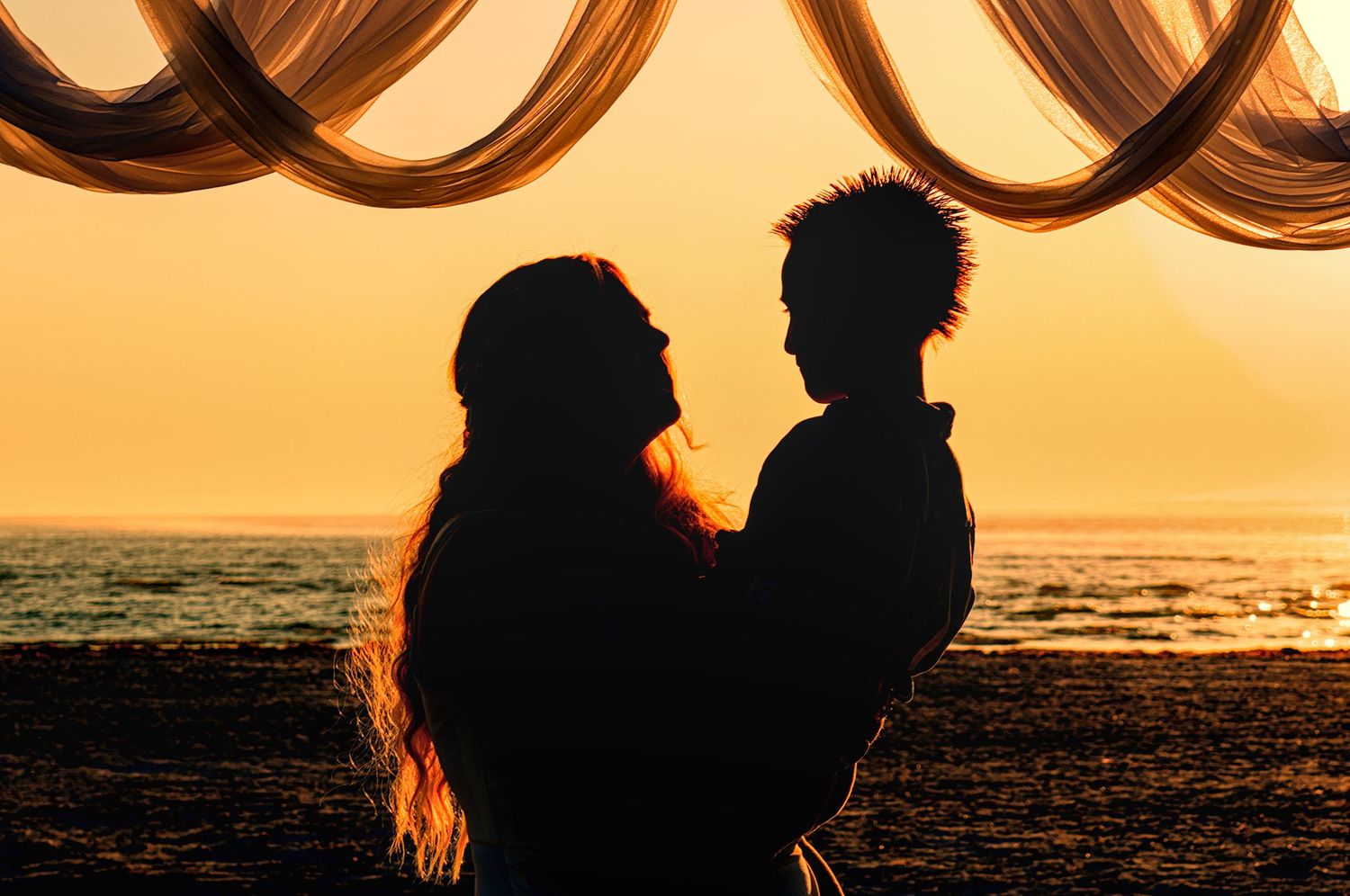 Silhouette of two people embracing on beach at sunset with draped fabric decor overhead.