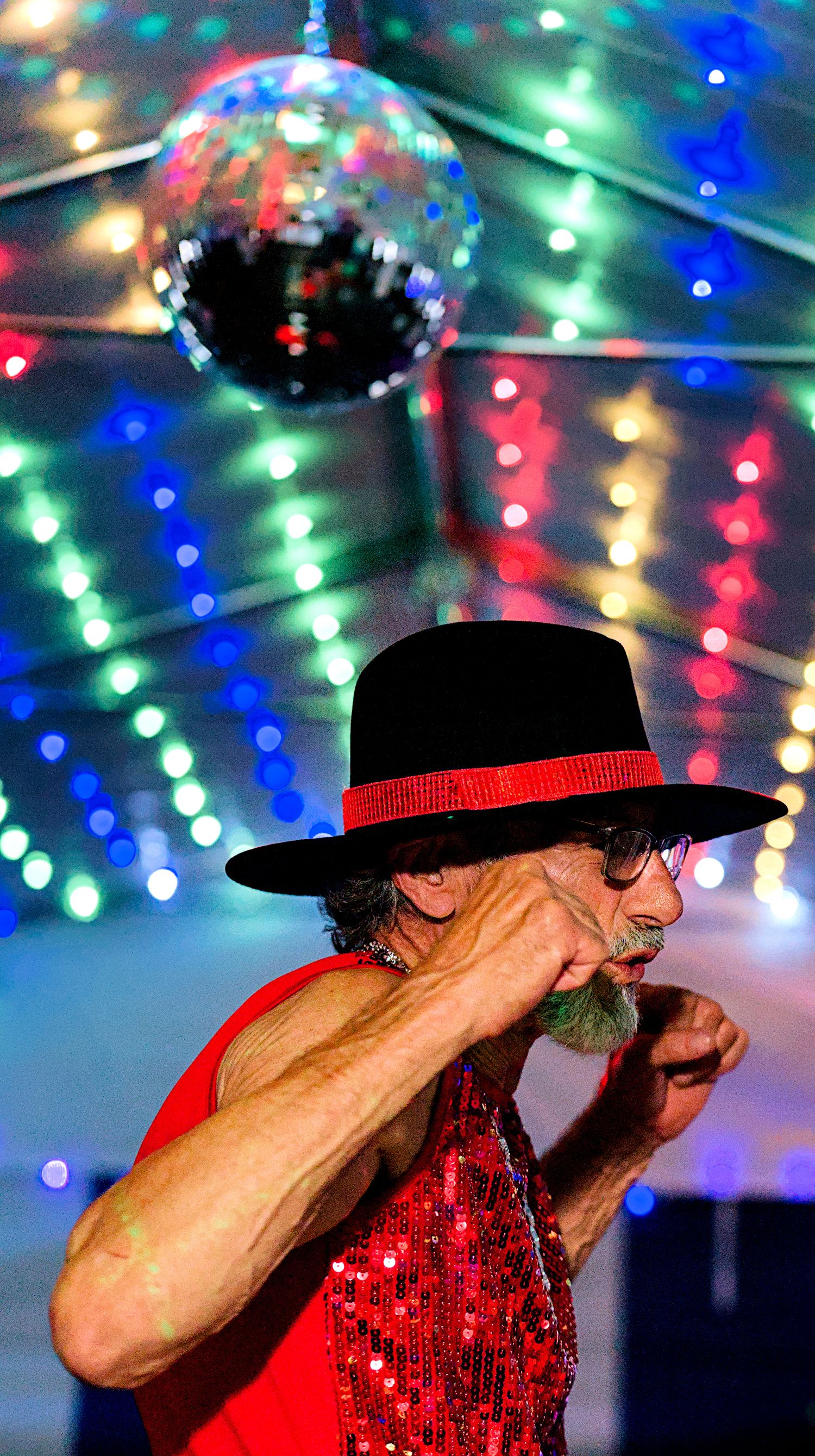 Elderly man dancing in red sequin vest and black hat under disco ball with colorful lights at party.