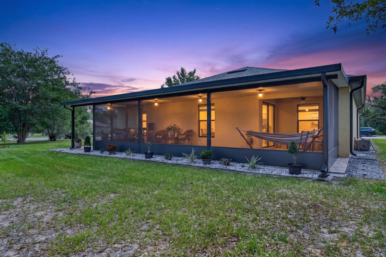 A screened porch glows warmly at twilight on a Florida home surrounded by lush green lawn under a colorful sunset sky.