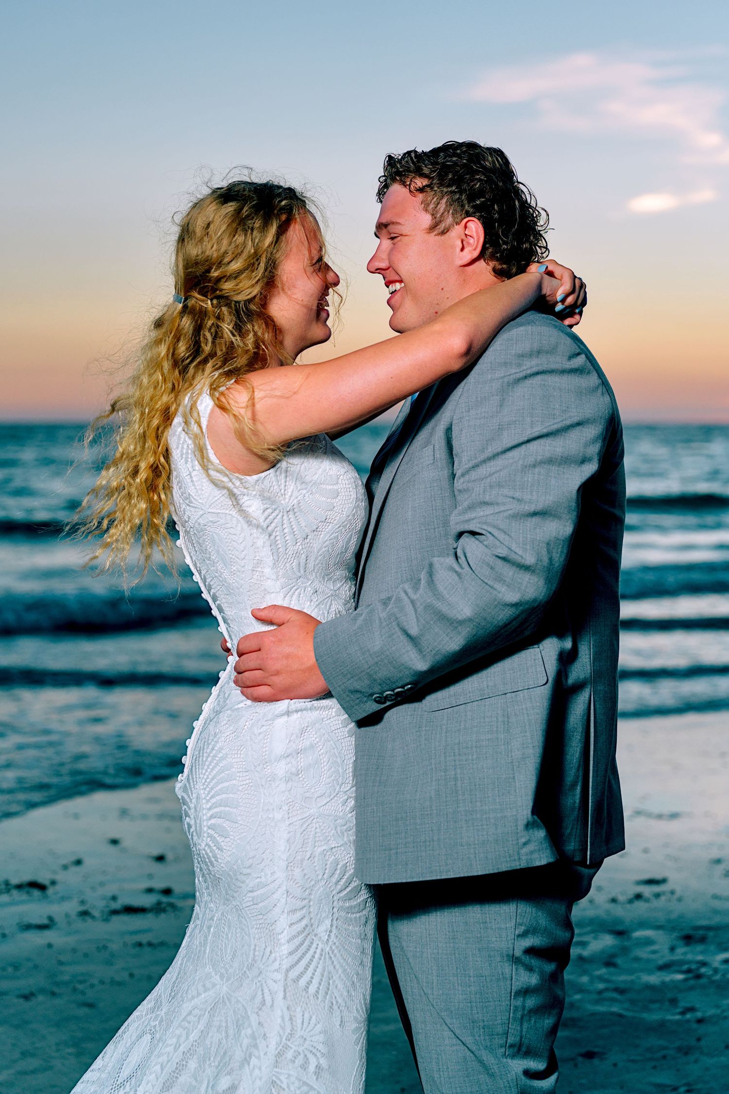Bride and groom embracing on beach at sunset, smiling joyfully in white gown and gray suit.