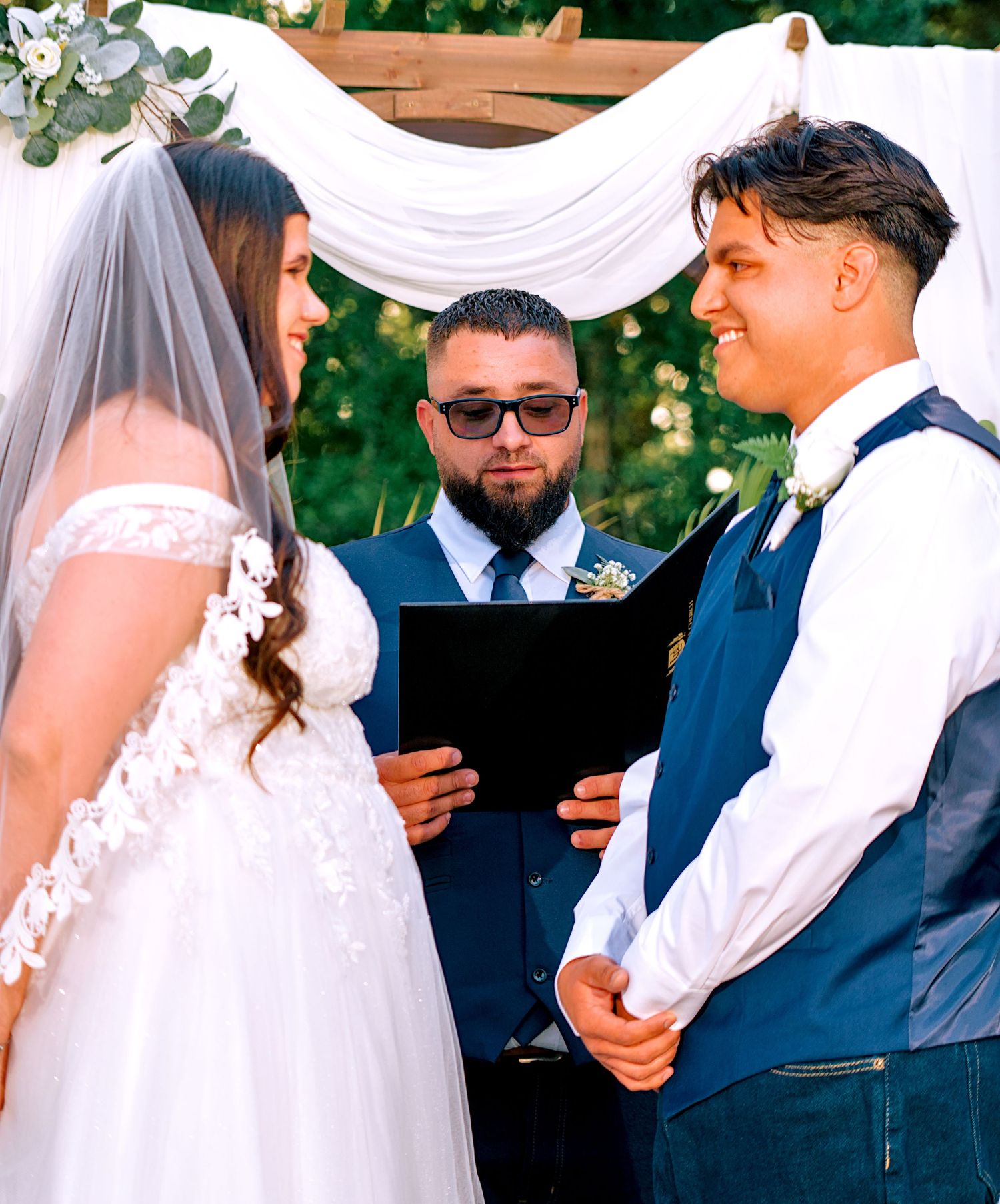 Bride and groom exchanging vows at outdoor wedding ceremony with officiant under floral arch.