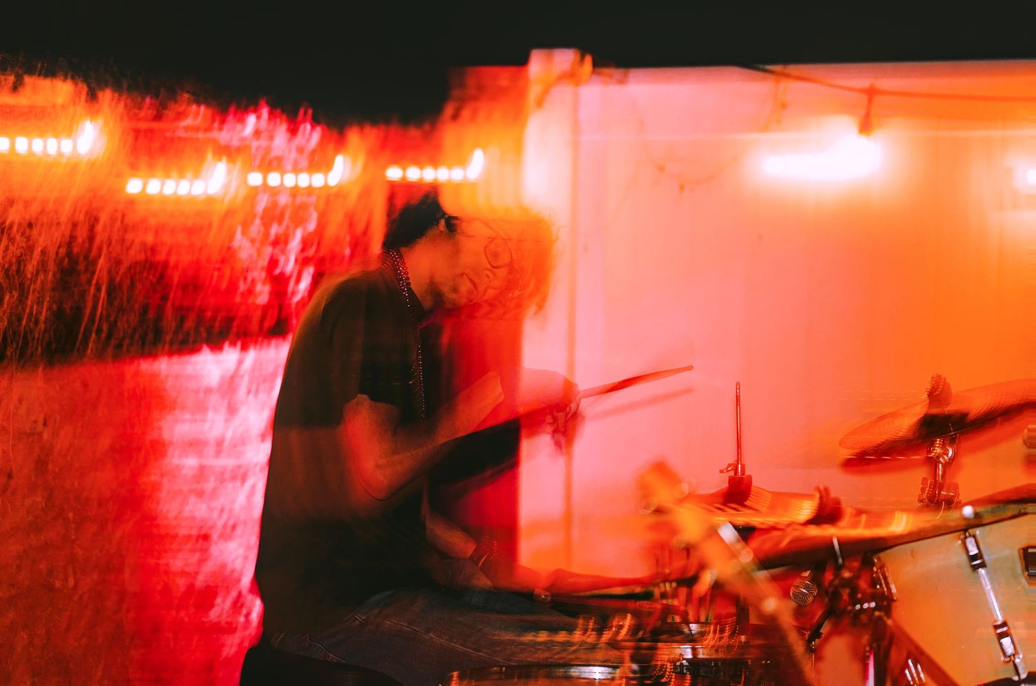 A drummer performs on stage bathed in dramatic red lighting during a live concert performance.