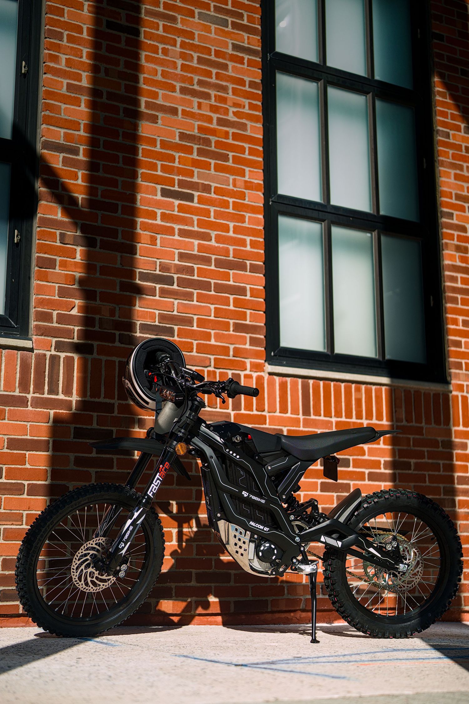 Black off-road dirt bike parked against a red brick building wall on a sunny day.