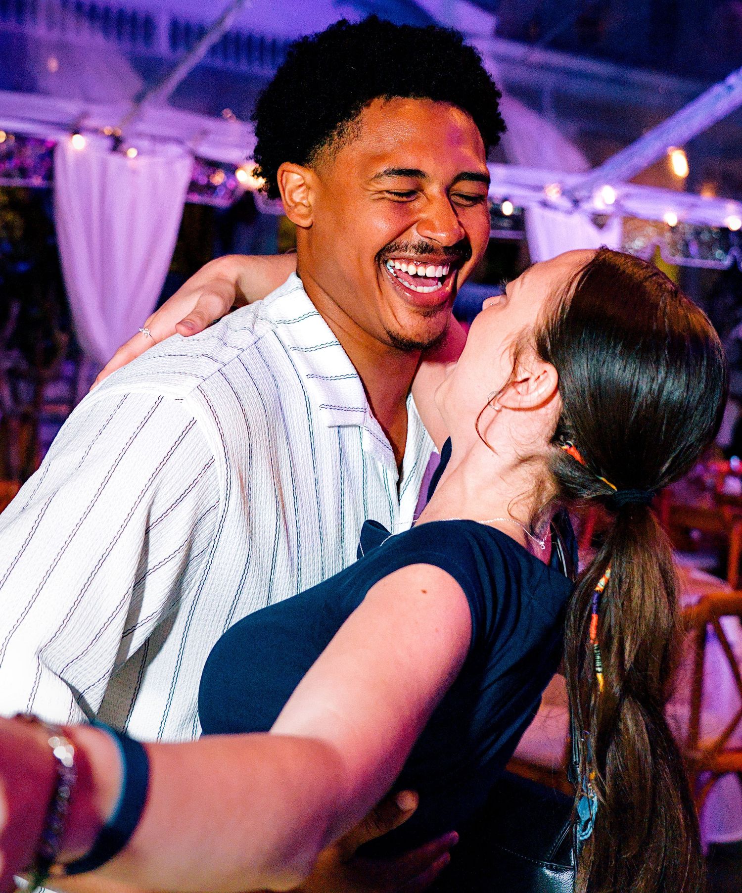 Two people laughing and embracing joyfully at an indoor event with string lights in the background.