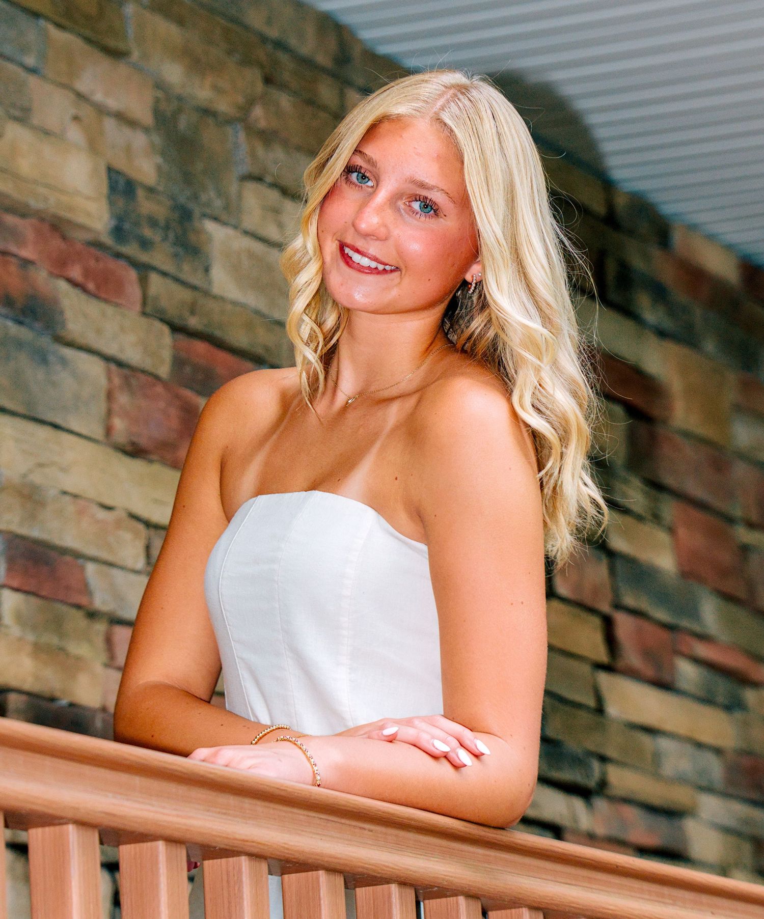 Smiling young blonde woman in white strapless dress leaning on wooden railing against stone wall backdrop.