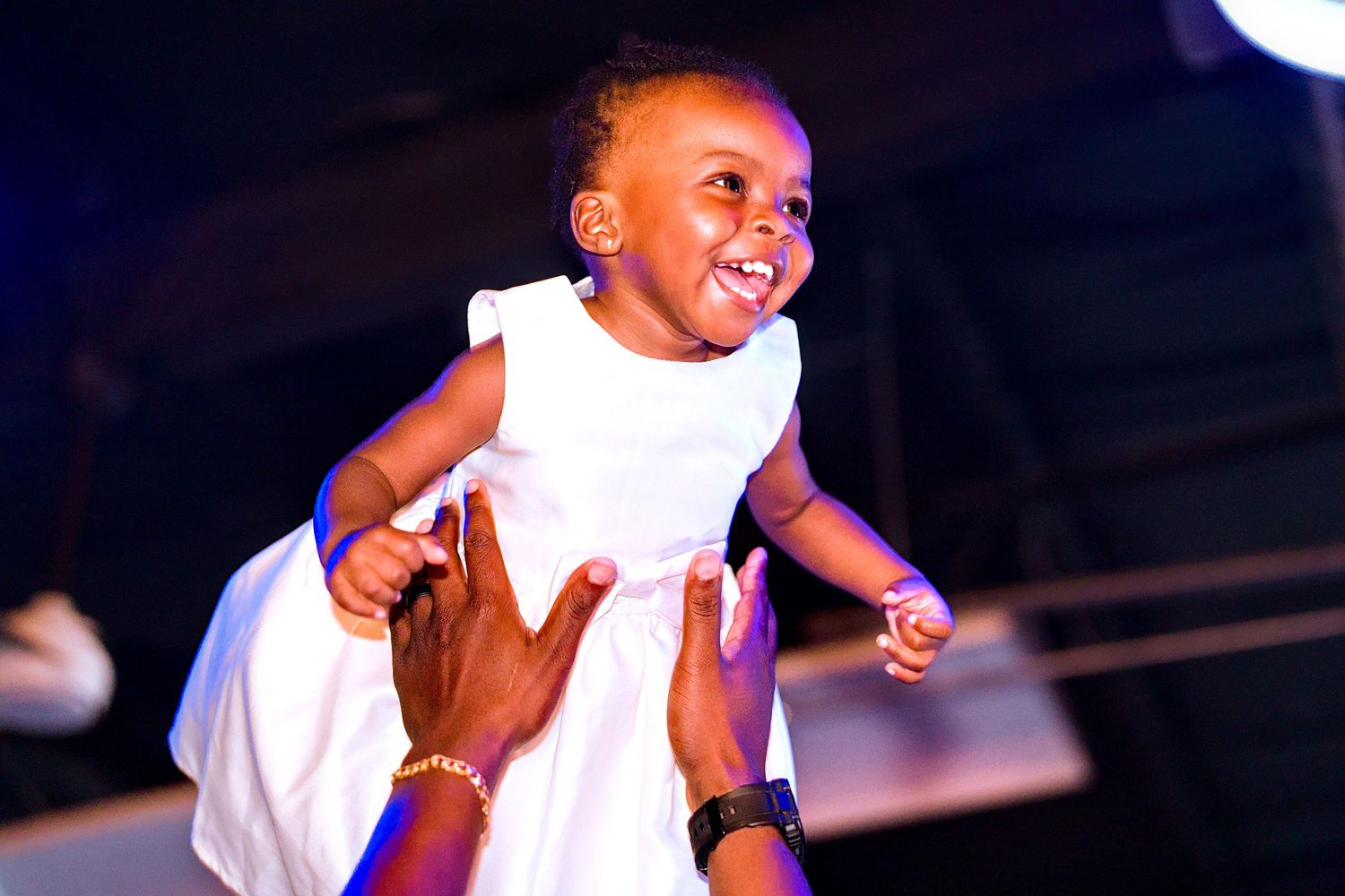 Joyful baby girl in white dress being lifted up by adult hands at a festive event.