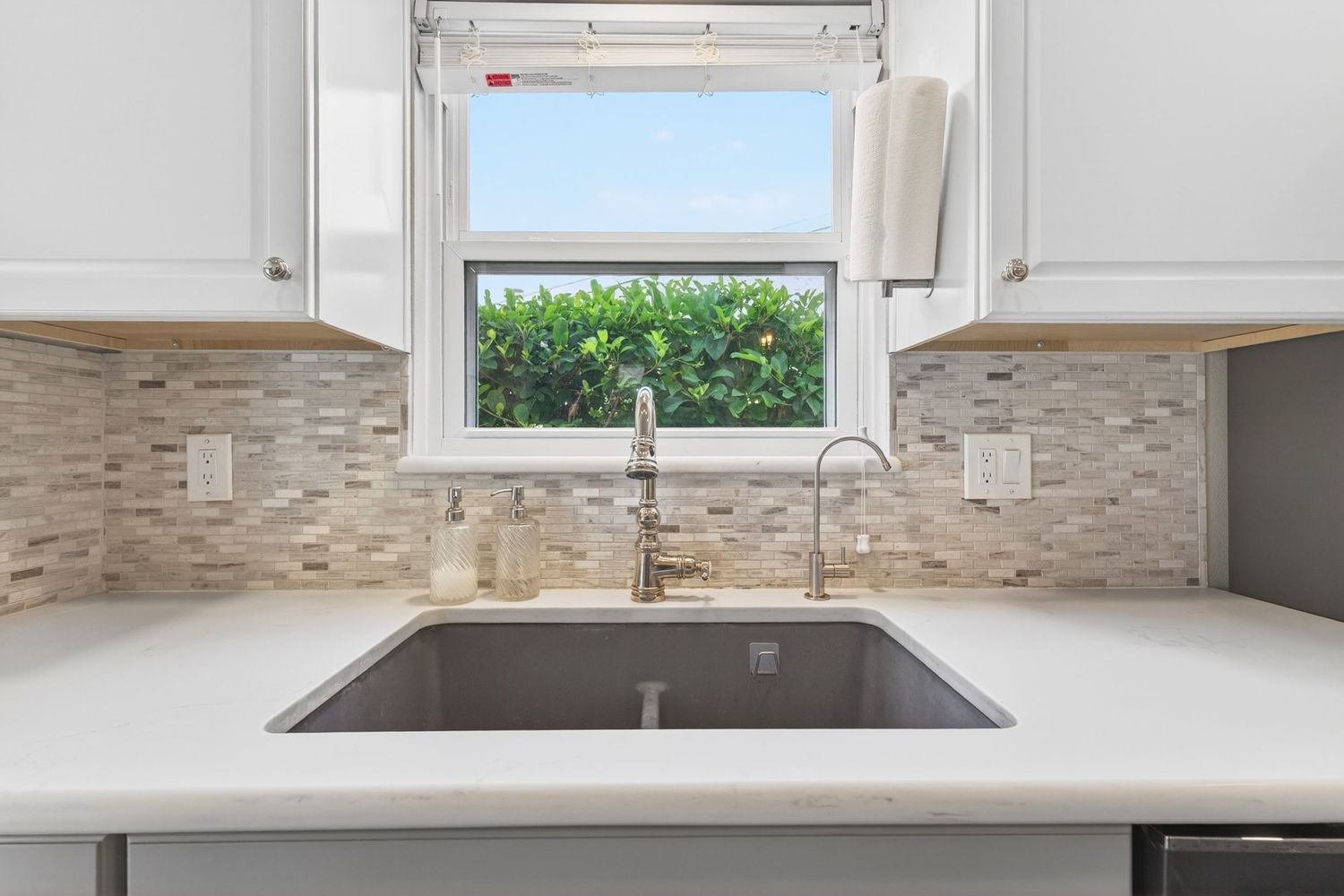Modern kitchen sink with mosaic tile backsplash, white cabinets, and a window overlooking lush greenery.