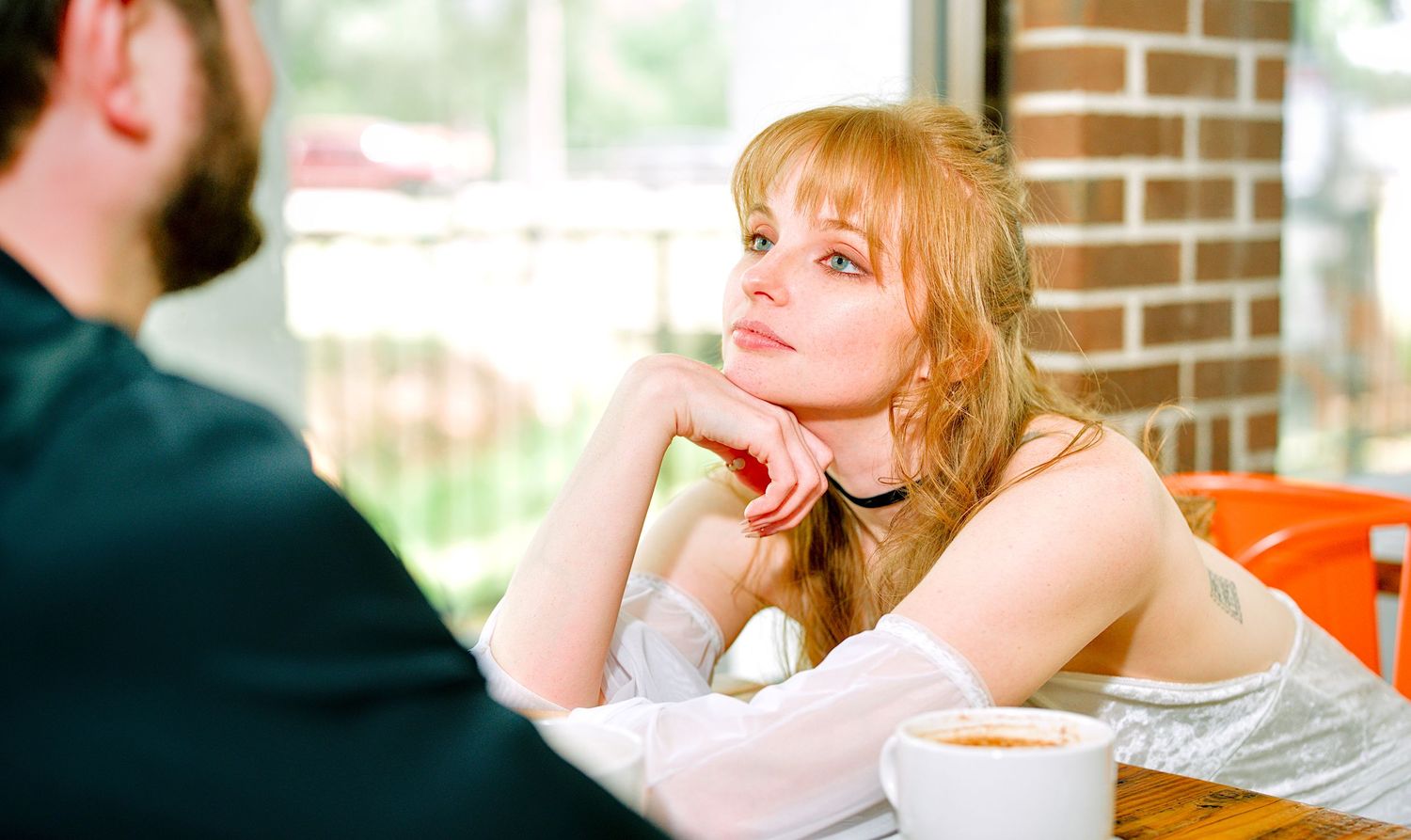 Elegant redhead woman in white dress gazing thoughtfully at a man in a cafe setting.