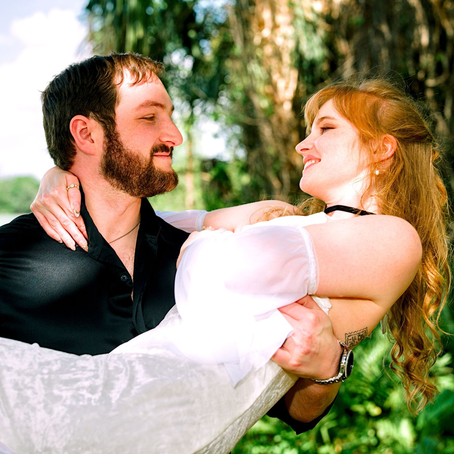 Bearded man in black vest carrying smiling redhead woman in white dress outdoors.
