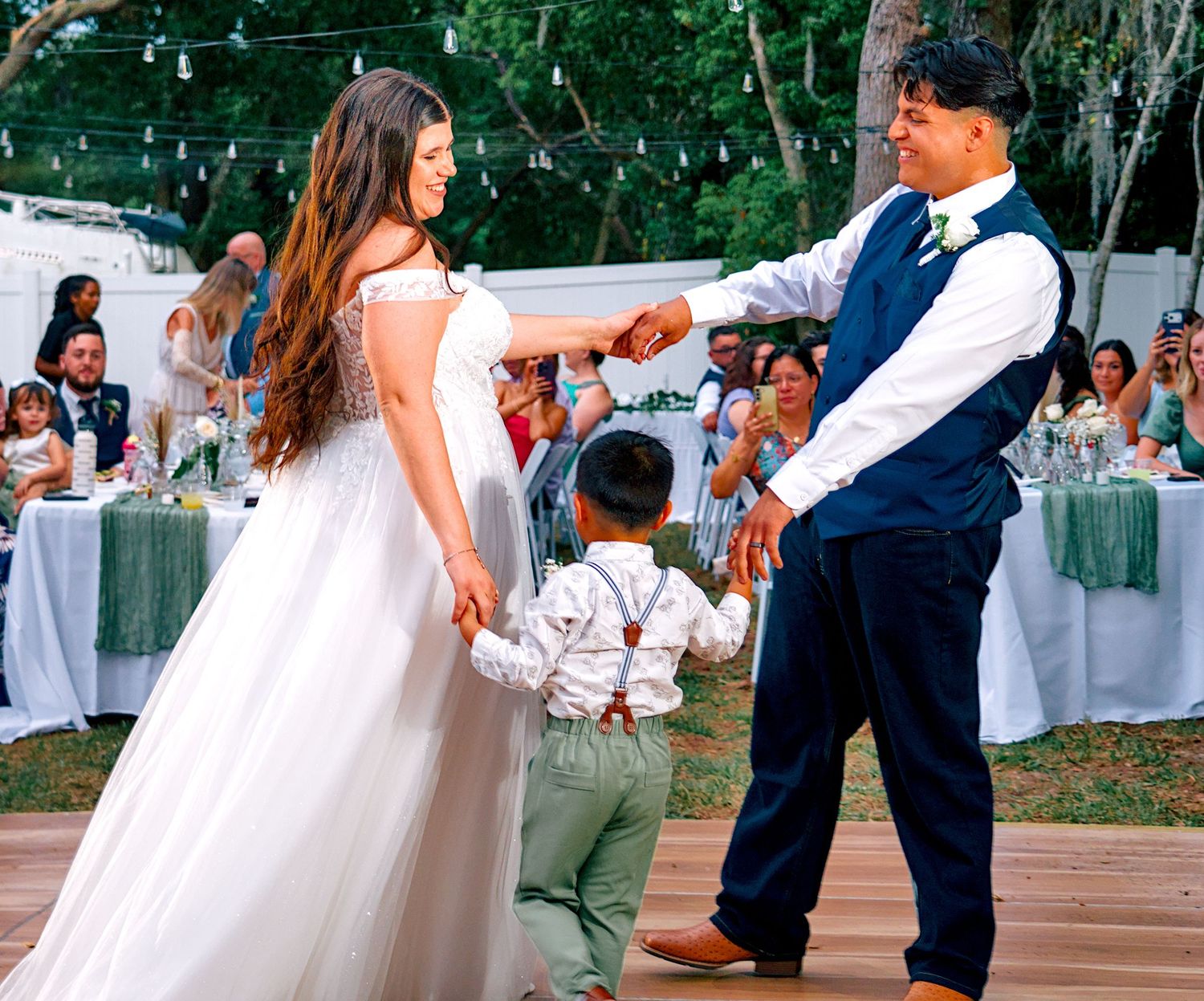 Bride and groom share first dance with young child on outdoor wedding reception dance floor.