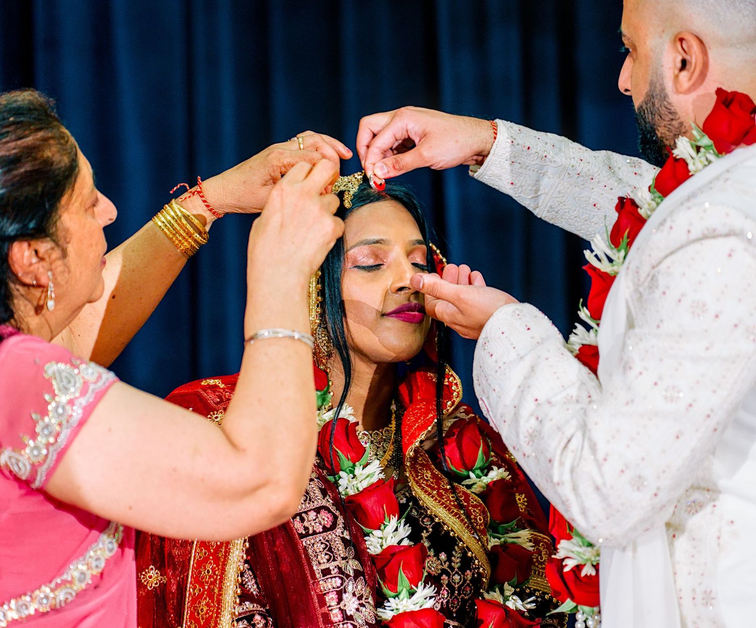Bride in red lehenga receiving floral hair adornment from groom and family.