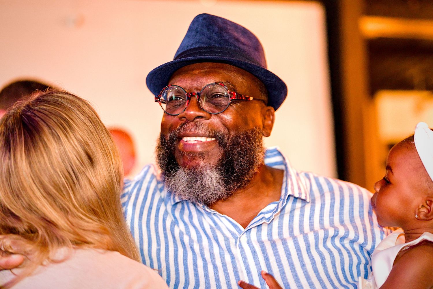 Smiling bearded man wearing a blue striped shirt and fedora hat at a celebration event.
