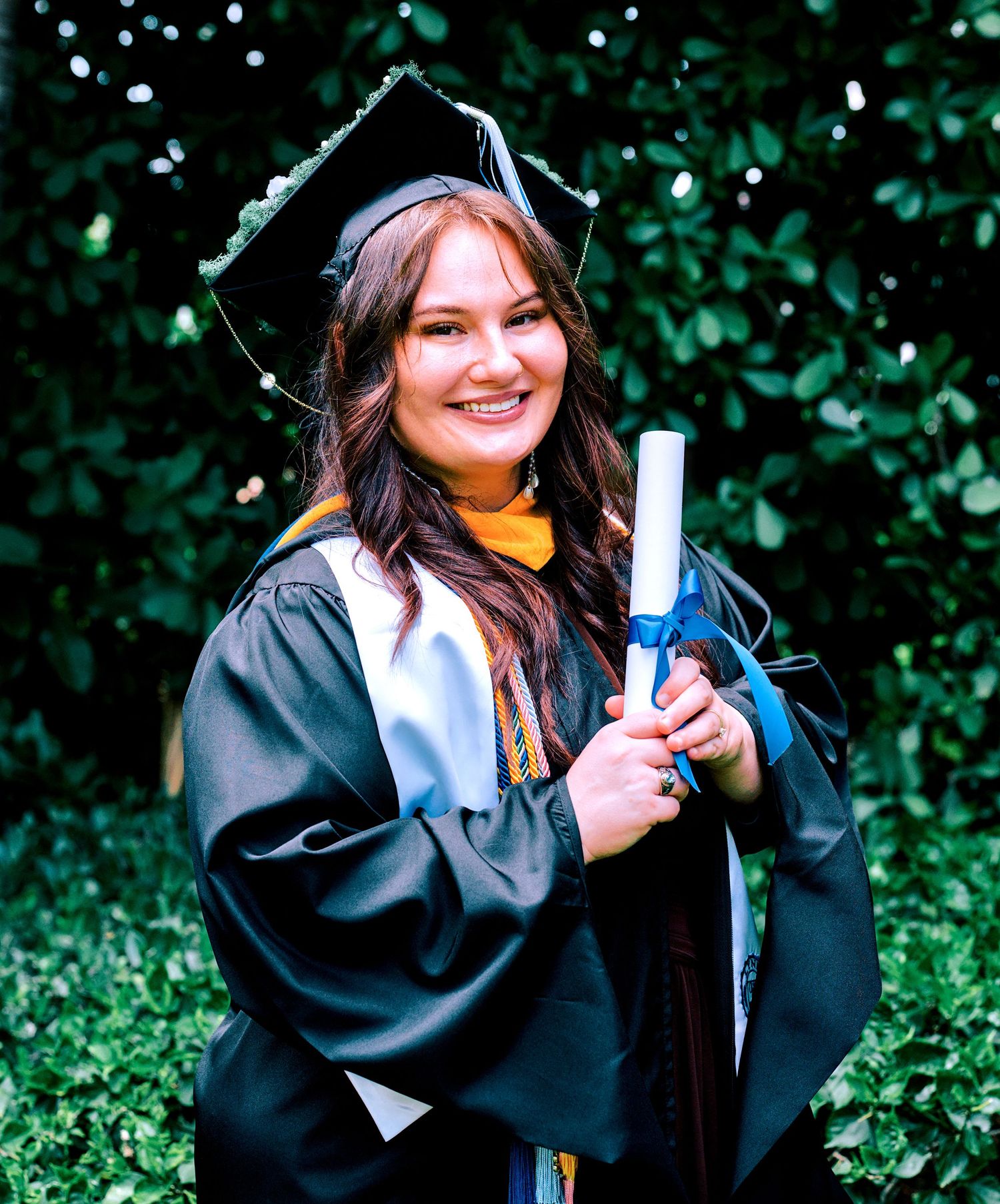Smiling graduate in black cap and gown holding diploma outdoors.