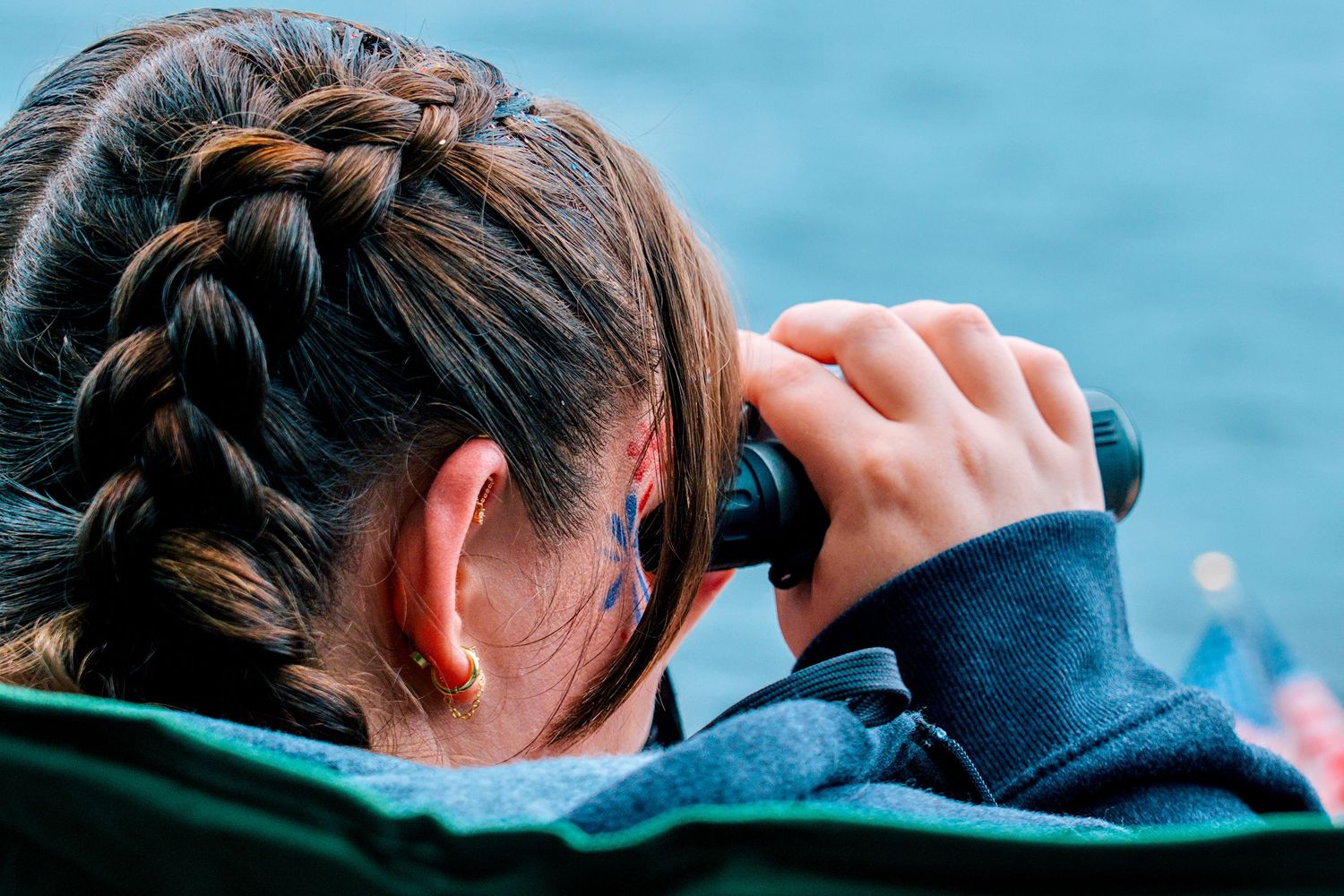 Young girl with braided hair looking through binoculars outdoors near water.