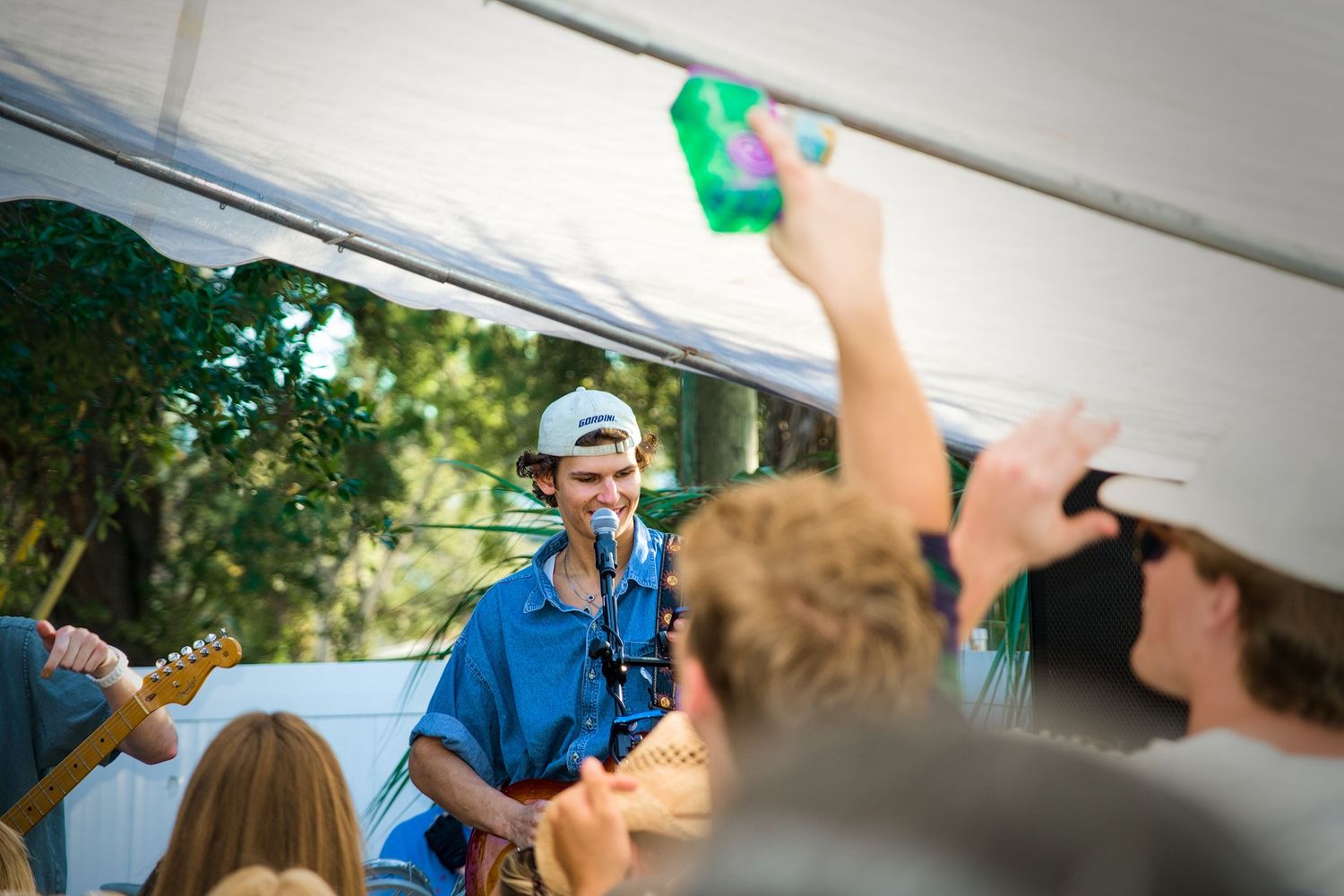 Young musician performs guitar under a tent at an outdoor festival while fans cheer and raise drinks in celebration.