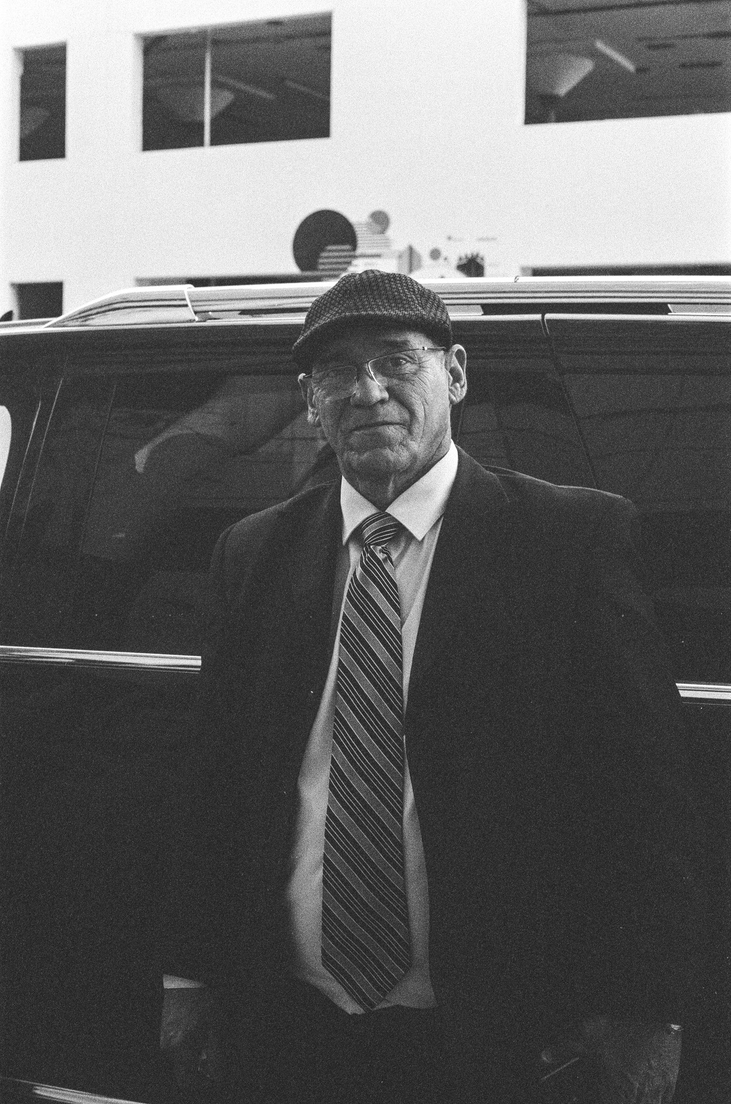 Elderly man in cap, glasses, and striped tie stands beside a black vehicle in a black and white photo.
