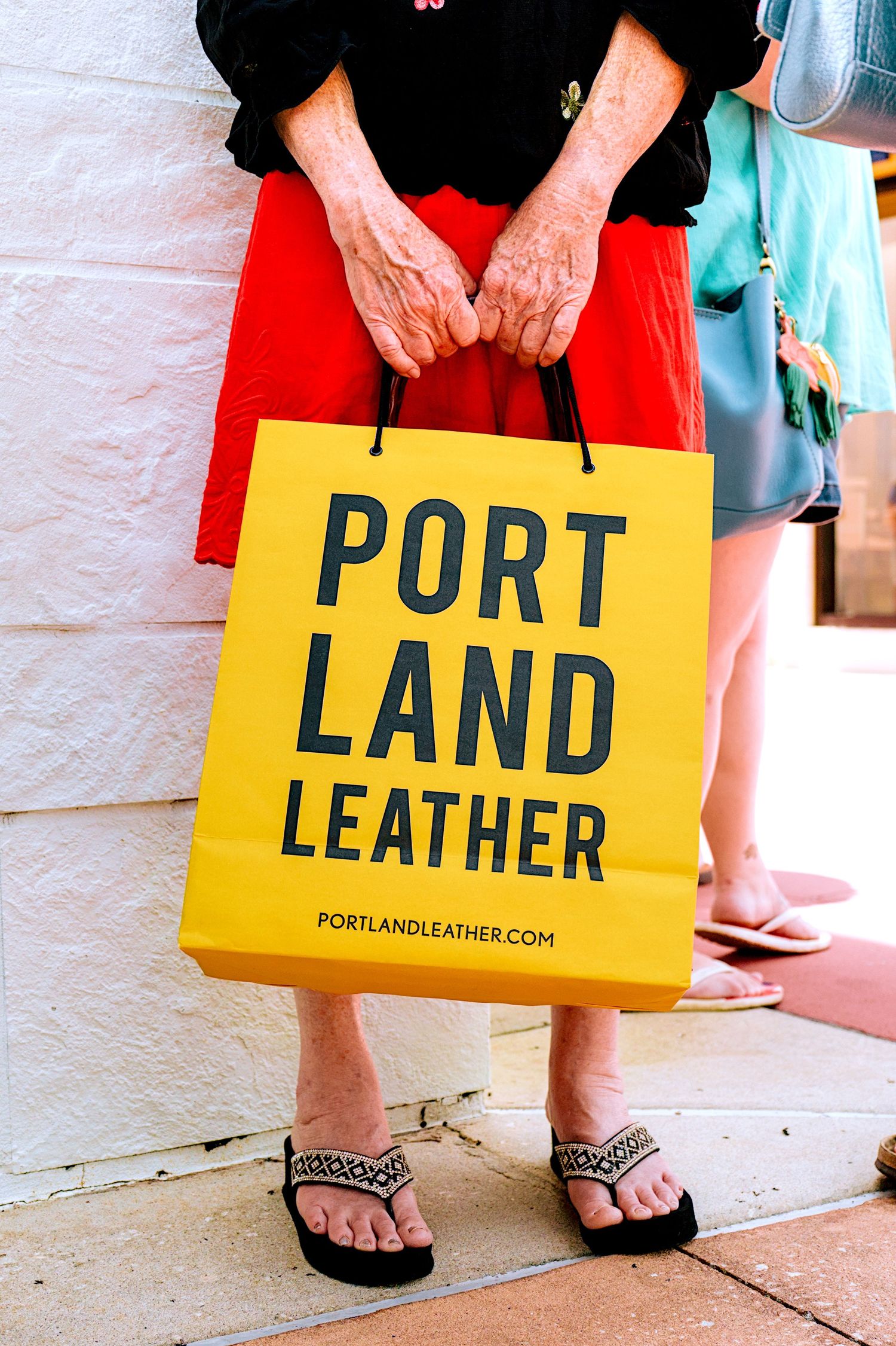 Person holding a yellow Portland Leather shopping bag while wearing flip flops and a red skirt outdoors.
