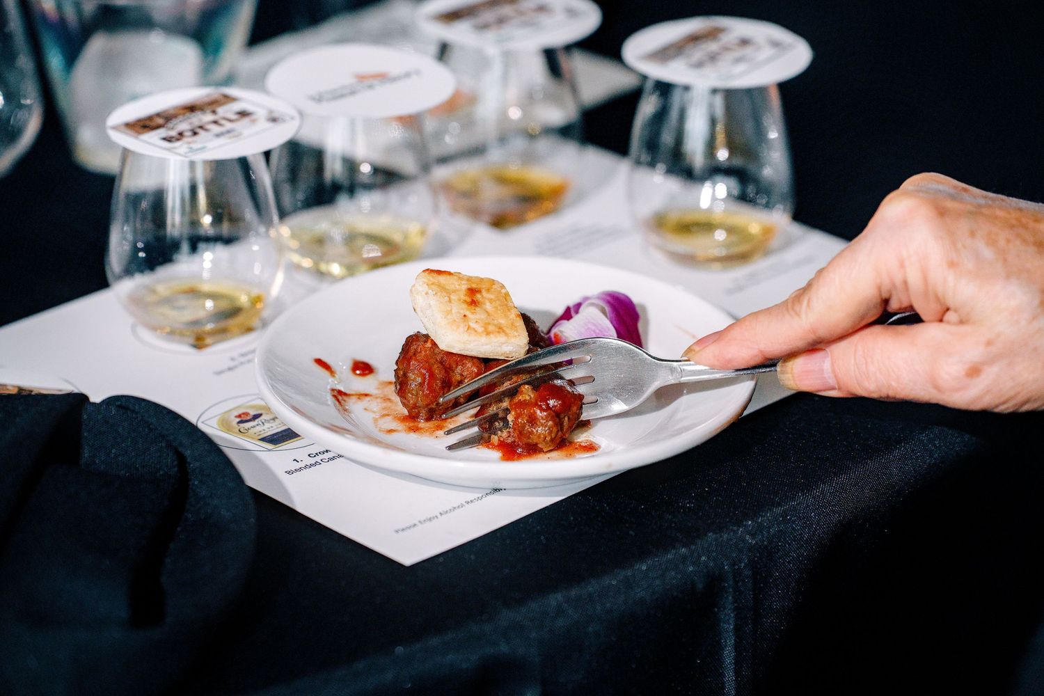 A hand holds a fork over a gourmet dish with wine glasses and tasting notes on a black tablecloth.