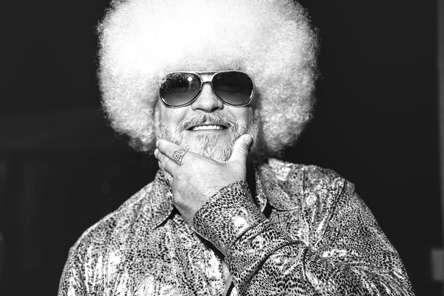 Man with white afro, sunglasses, and patterned shirt posing confidently in a black and white retro photo.