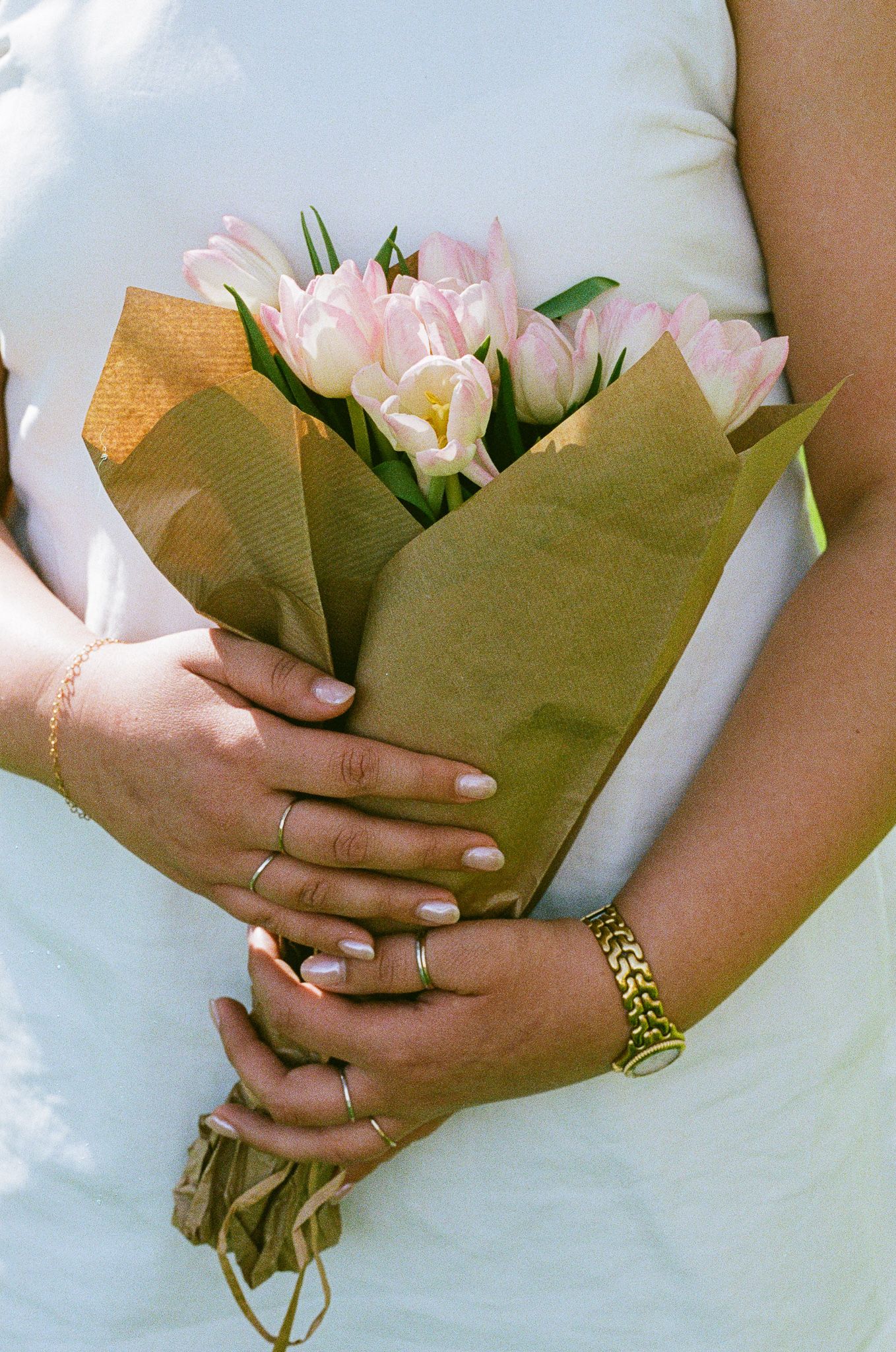 Close-up of hands holding a bouquet of pink tulips wrapped in brown kraft paper with gold jewelry accents.
