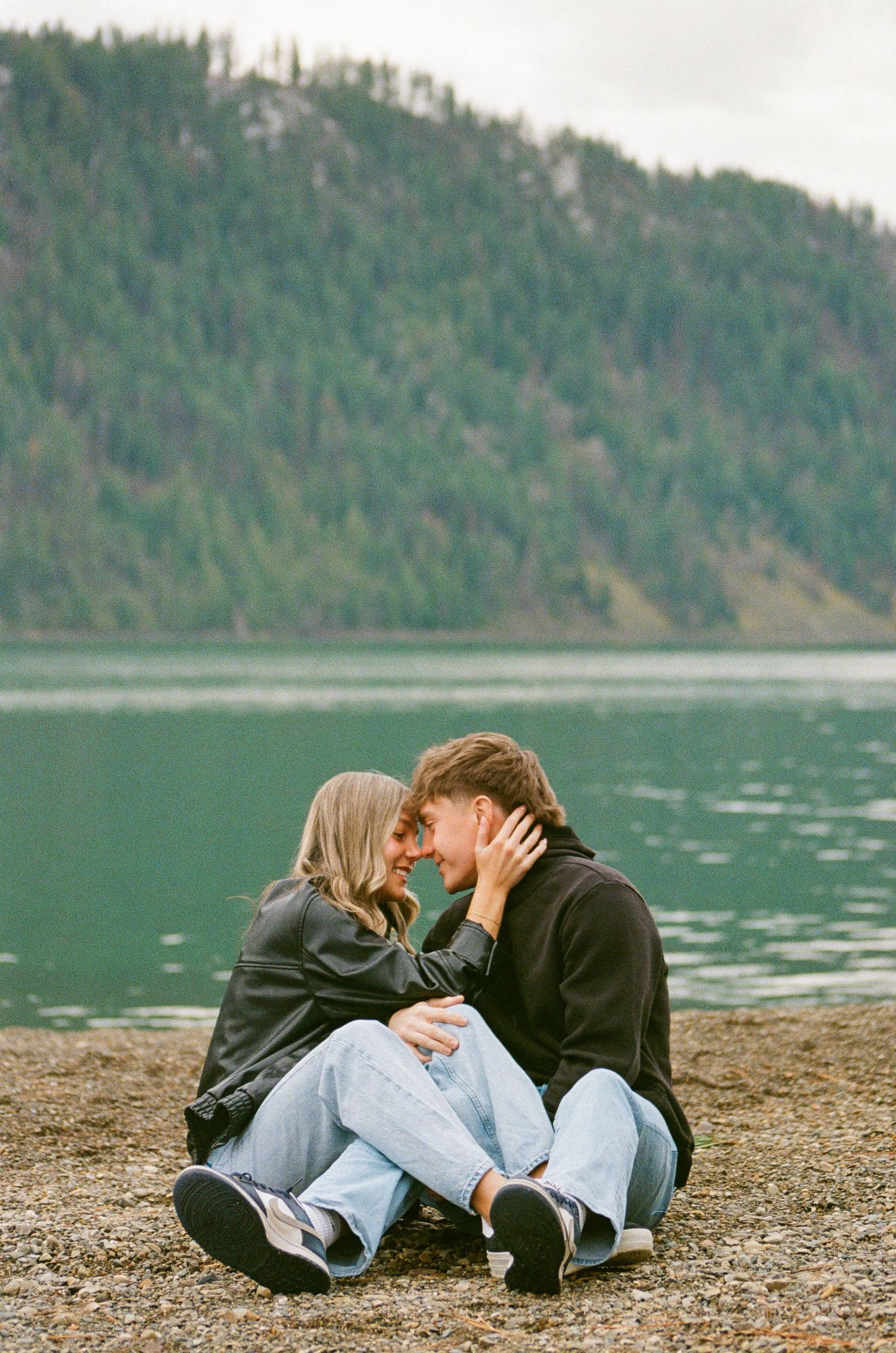 A couple sits closely by a serene mountain lake, sharing a romantic moment in matching black hoodies and jeans.