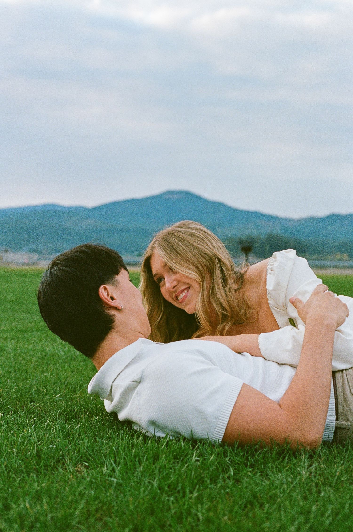 A couple laughing and embracing while lying on green grass with mountain scenery in the background.