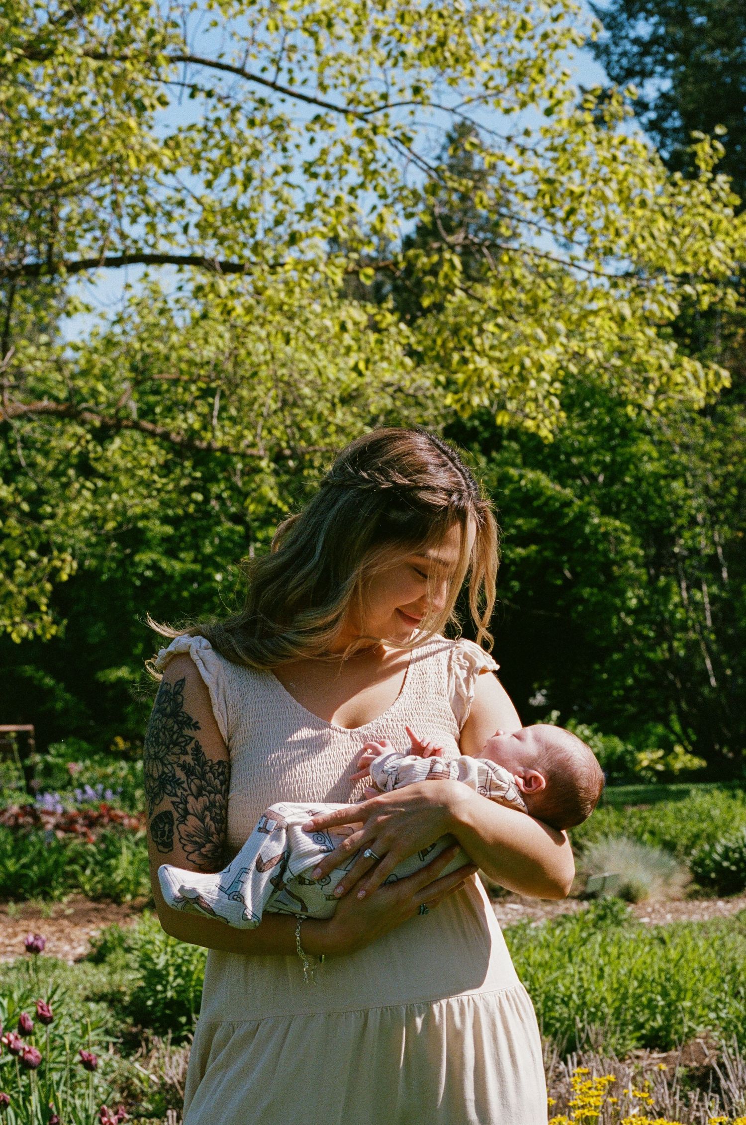 A tattooed mother in a cream dress cradles her newborn baby outdoors in a lush green garden on a sunny day.