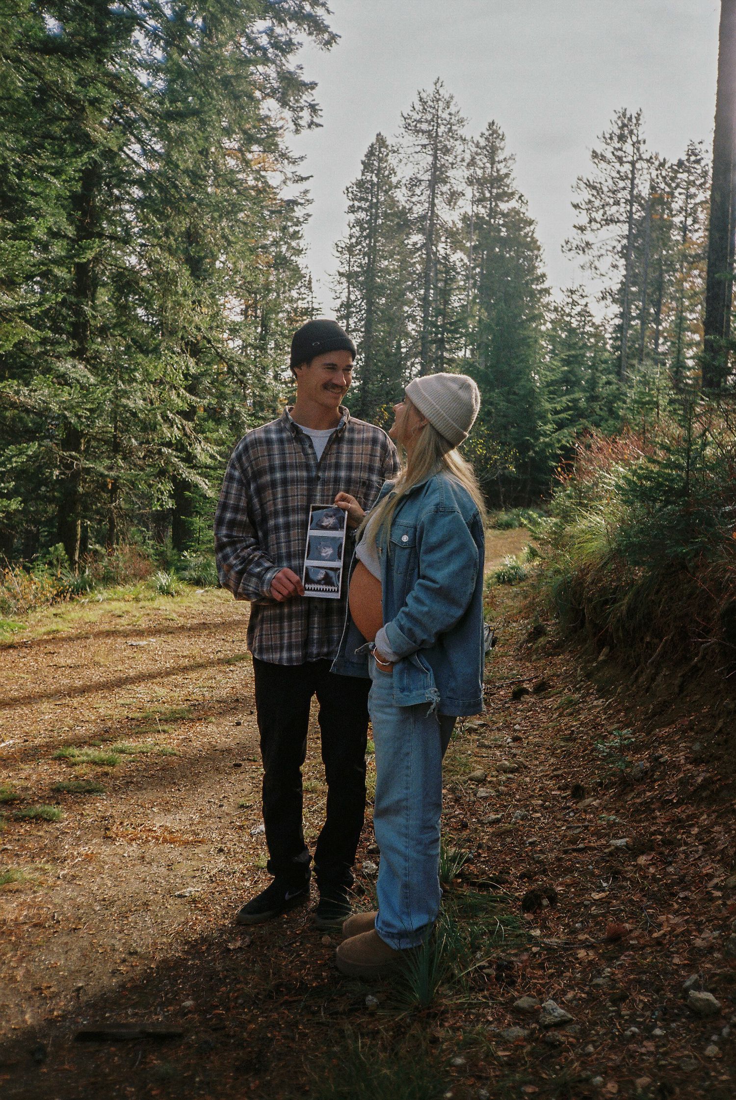 Couple holding ultrasound photo in a forest setting, announcing pregnancy on a woodland trail.