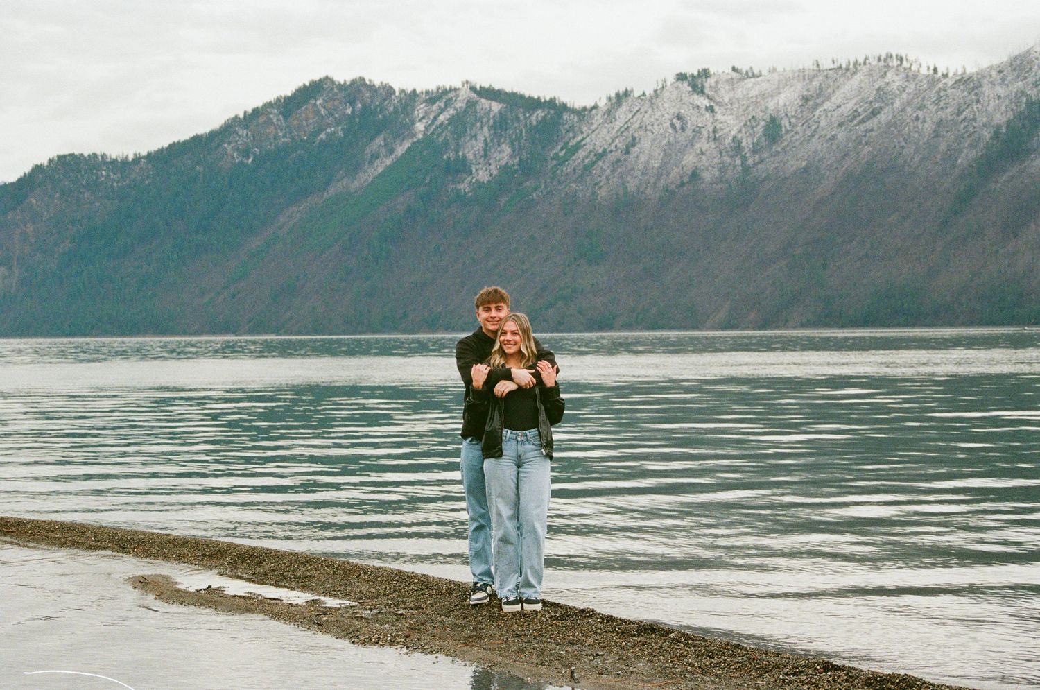 Couple embracing on a rocky shoreline with a scenic mountain lake backdrop on a cloudy day.