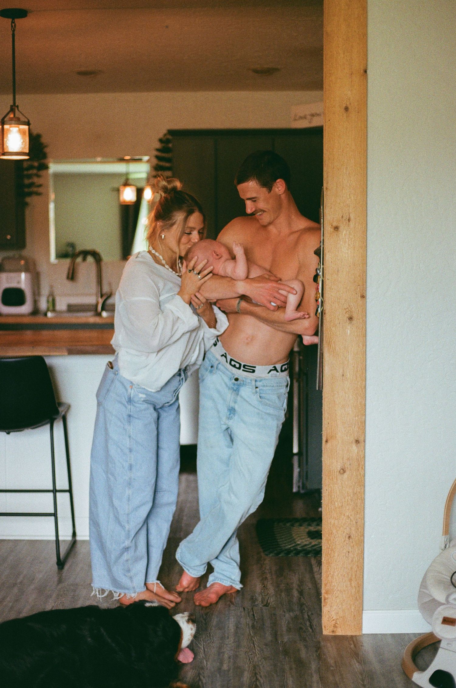 A couple in jeans holds their newborn baby together in a cozy home interior.