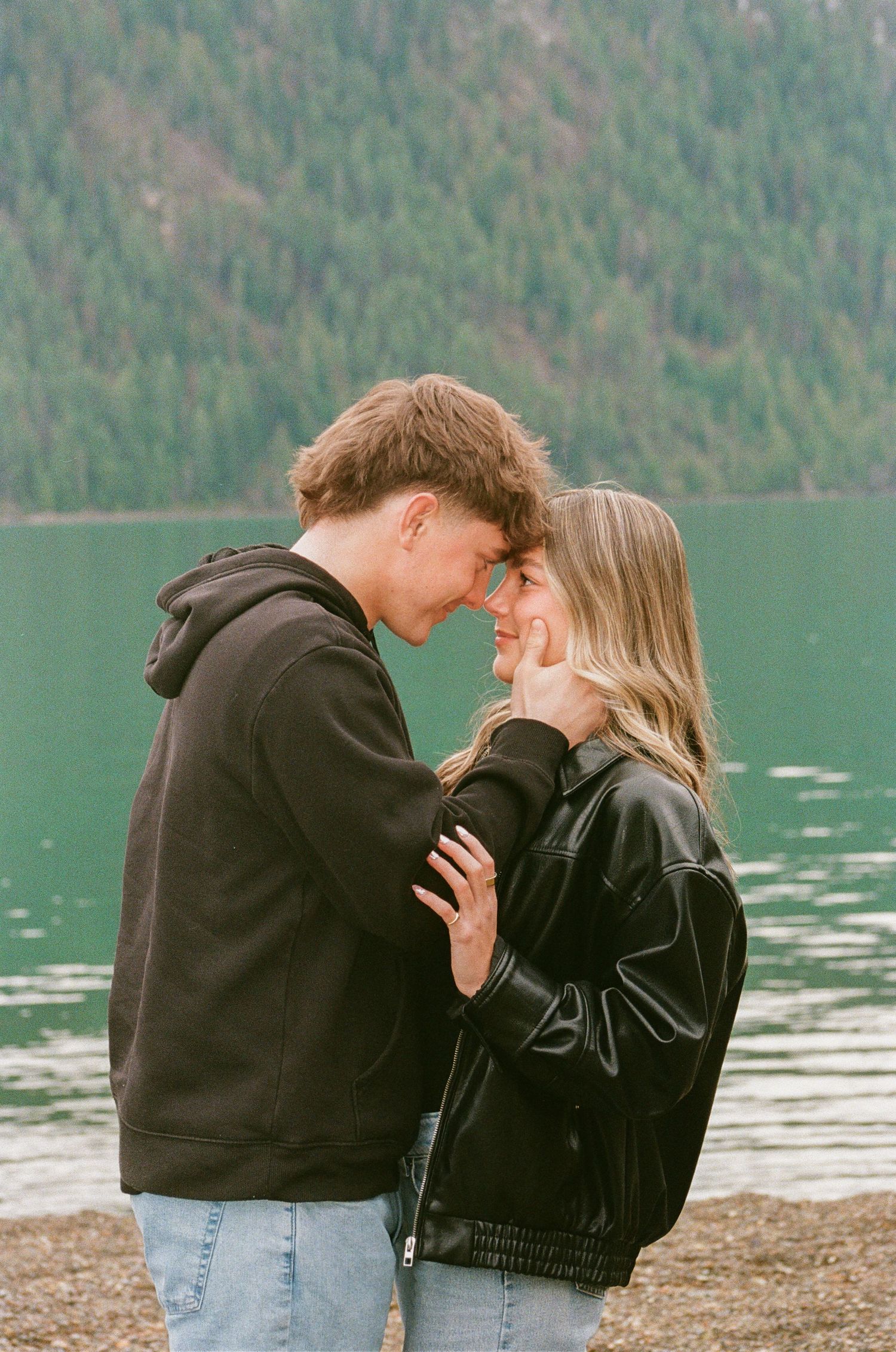 Couple sharing a romantic forehead-to-forehead moment by a serene mountain lake, wearing casual jackets.