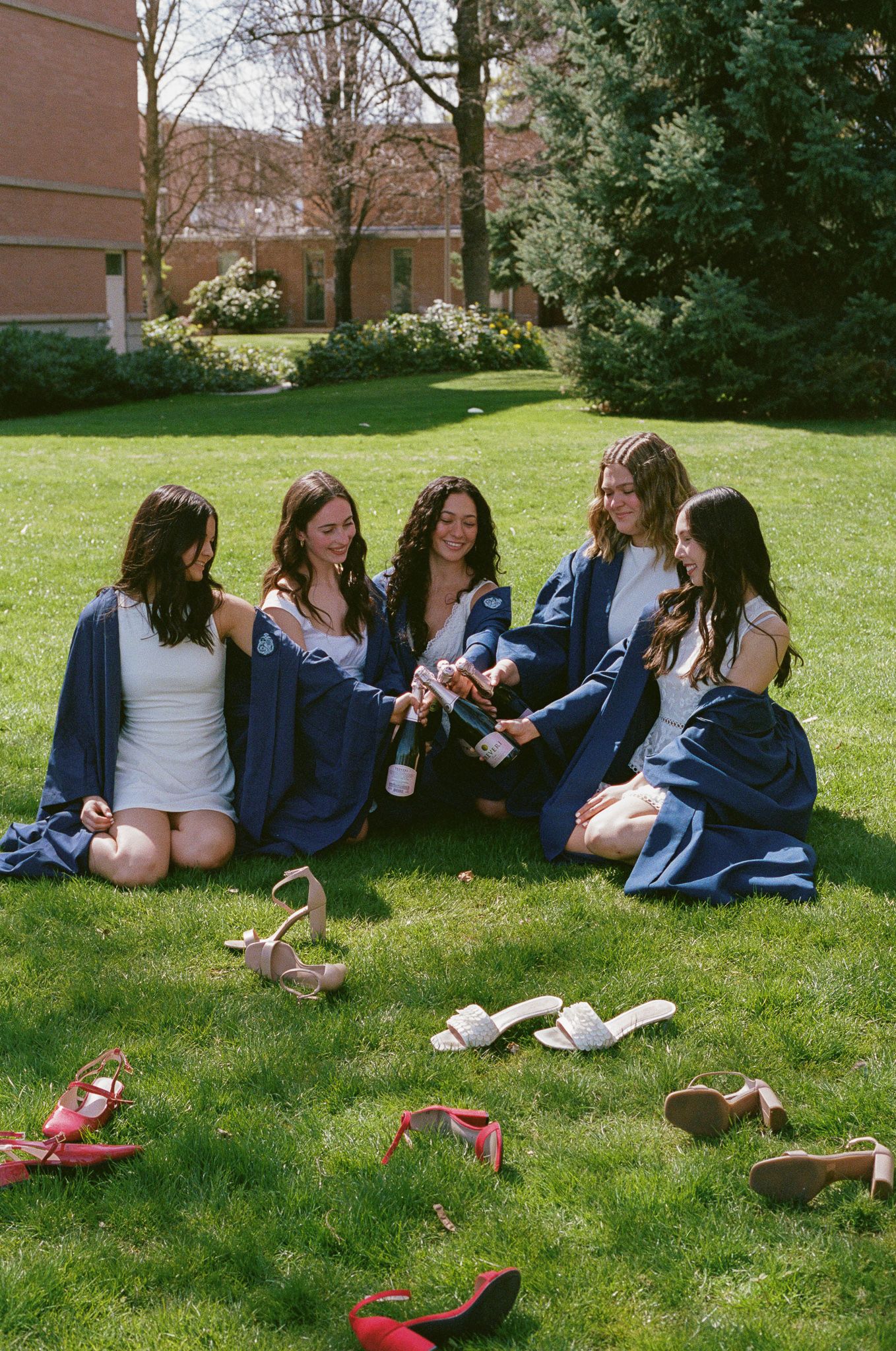 Five female graduates in navy robes sit on grass, celebrating with champagne, shoes scattered around them.