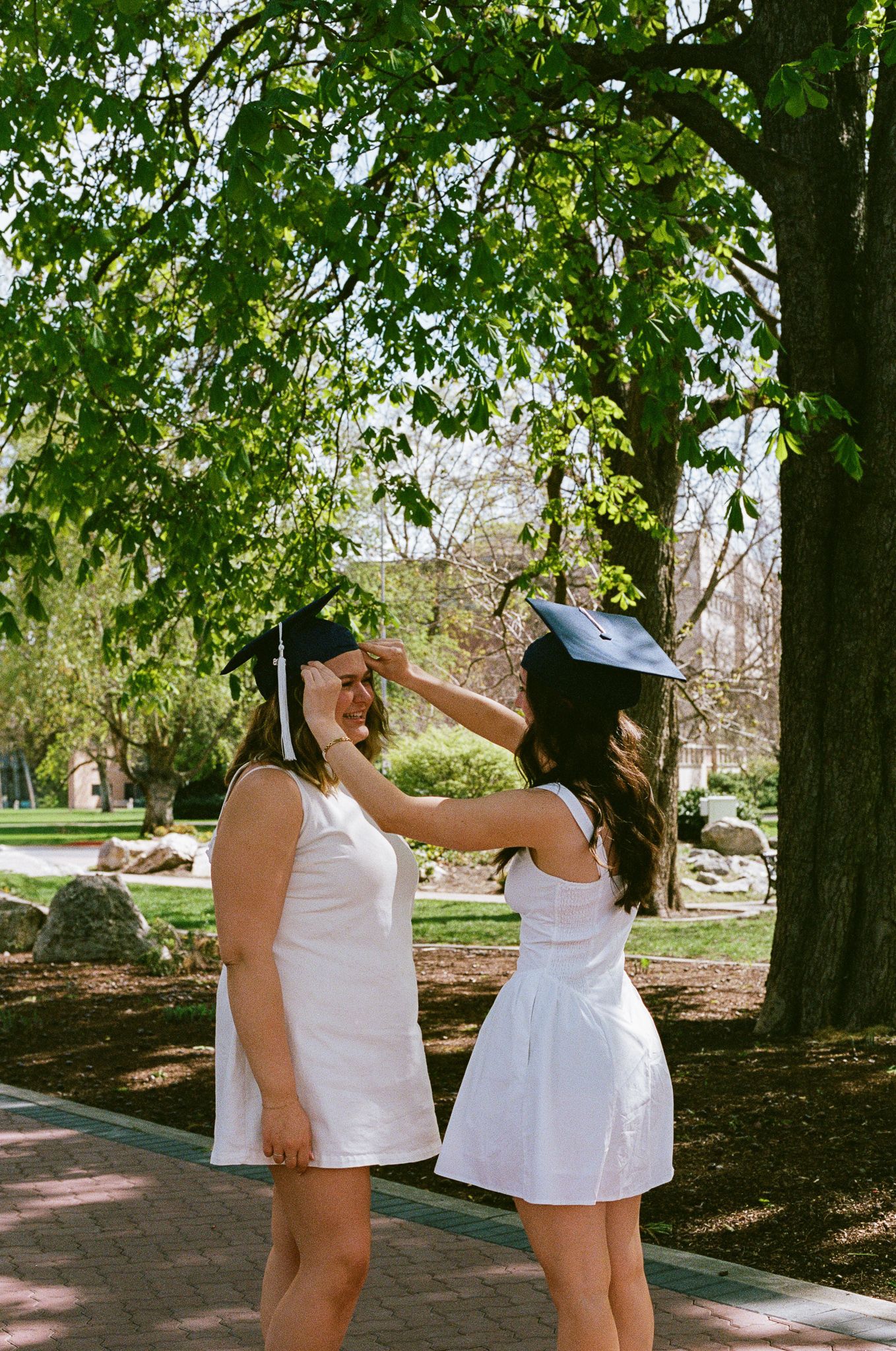 Two female graduates in white dresses exchange graduation caps outdoors under green trees on a sunny day.