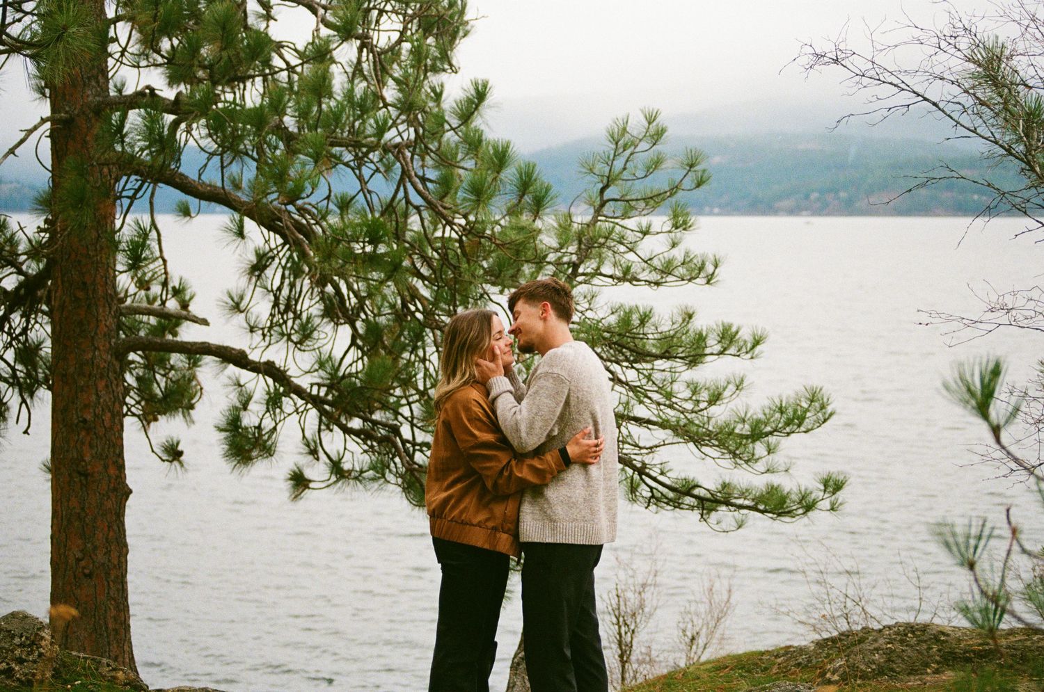 Couple embracing romantically by a lakeside pine tree with misty mountains in the background.