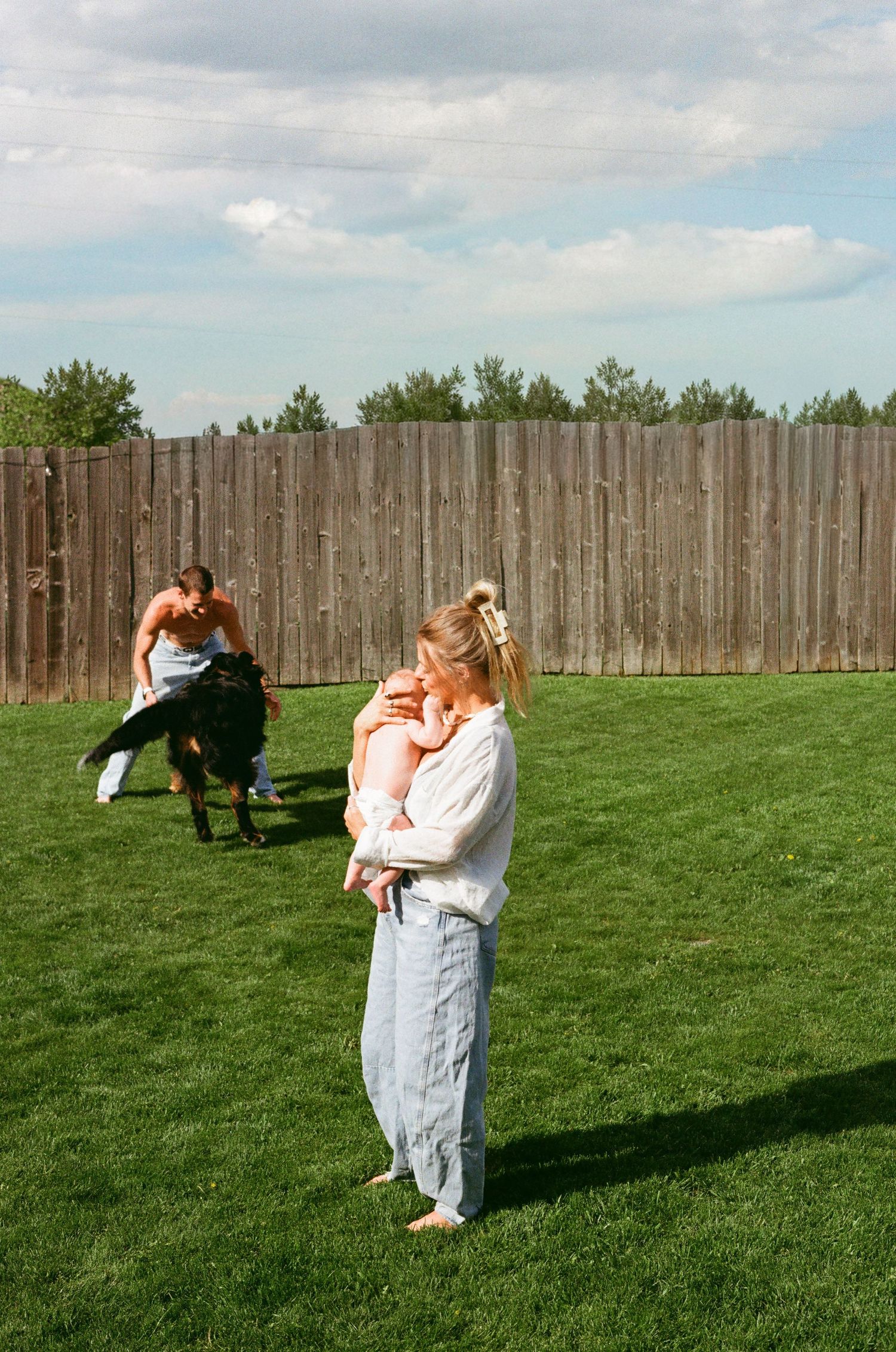 A woman holds a baby in a backyard while a shirtless man plays with a child on green grass near a wooden fence.