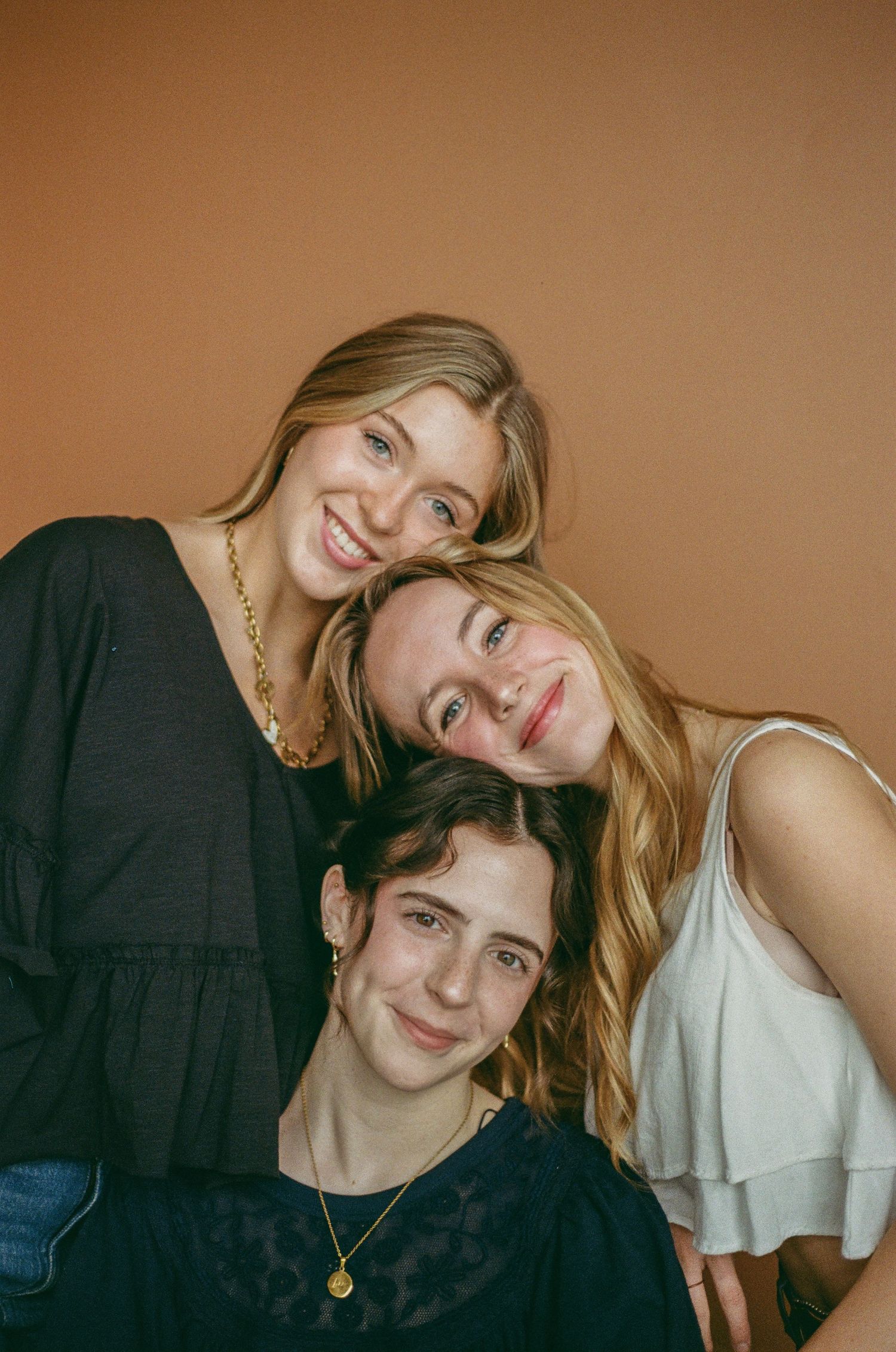 Three smiling young women posing together against a warm brown background in a cozy portrait photo.