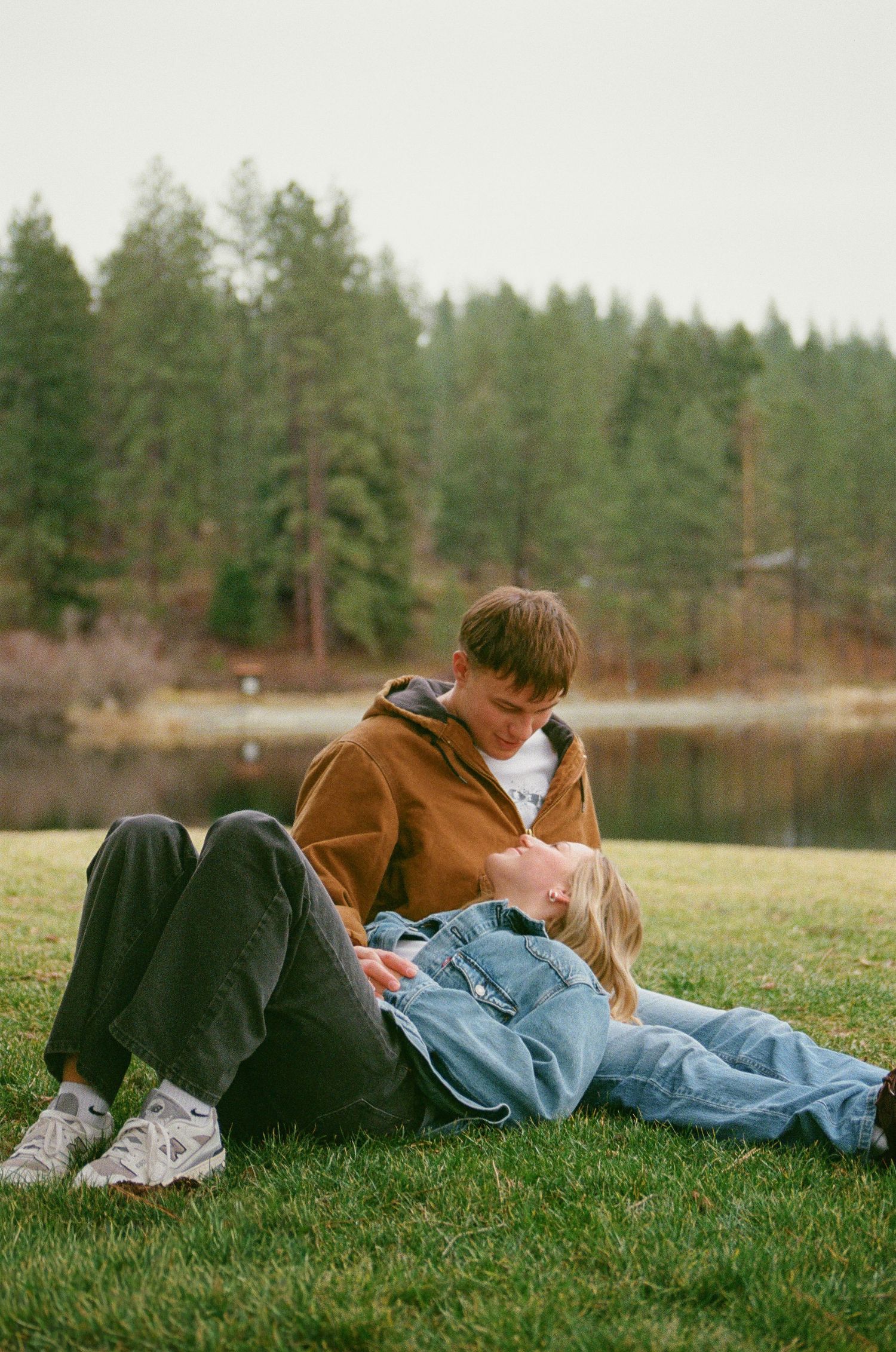 A young couple sits on green grass near a lake, surrounded by pine trees, sharing a tender romantic moment outdoors.